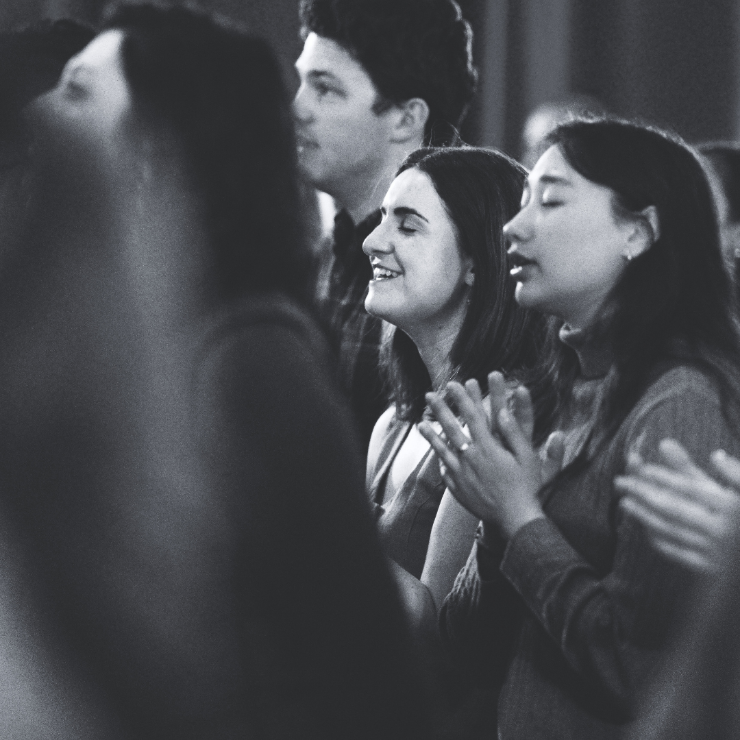 A group of people praying with their eyes closed, hands clasped, and smiling, in a black and white photo taken at Grace Capital City church worship service.