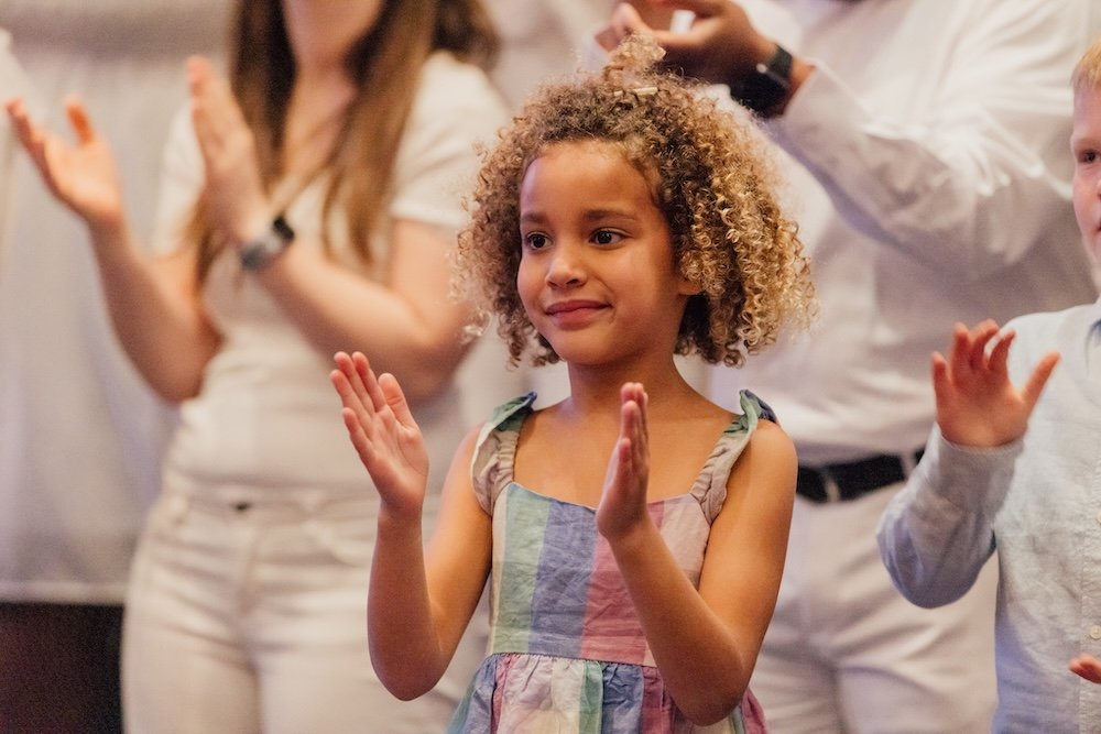 A young girl with curly hair claps her hands during a Grace Capital City church worship service in Washington, DC, with adults in the background also clapping.