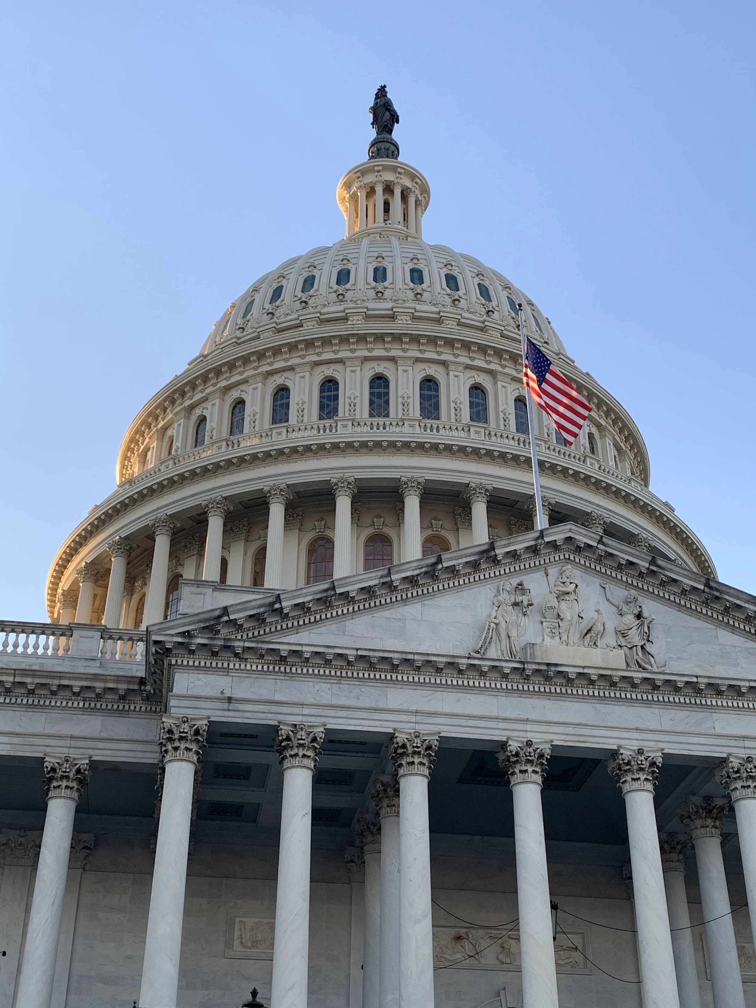 Front view of the United States Capitol in Washington, DC where Grace Capital City church has services. The view includes the iconic dome of the building with a blue sky background, American flag, and detailed white marble facade.