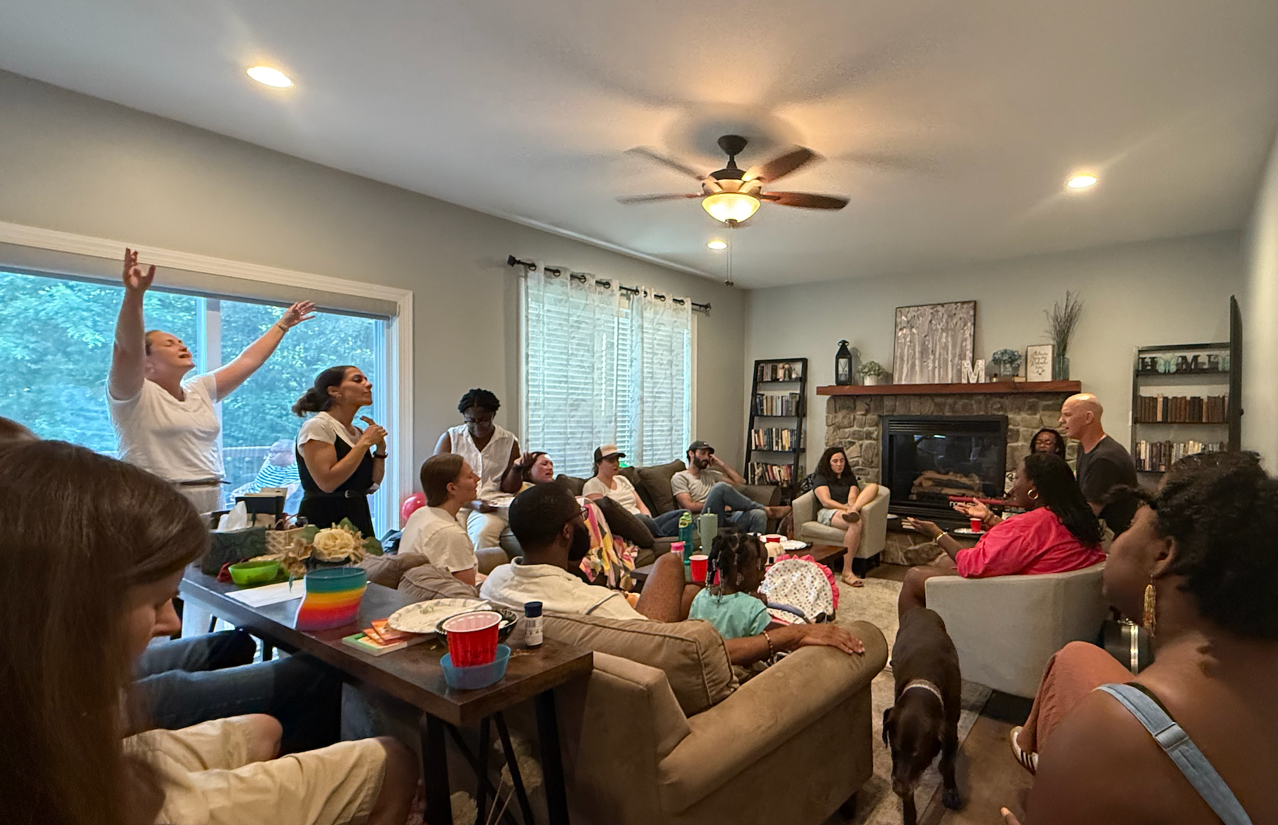 A group of people gathered in a living room, some sitting on couches and chairs, others standing, participating in a member social gathering for Grace Capital City church in Washington, DC, with a fireplace and bookshelves in the background.