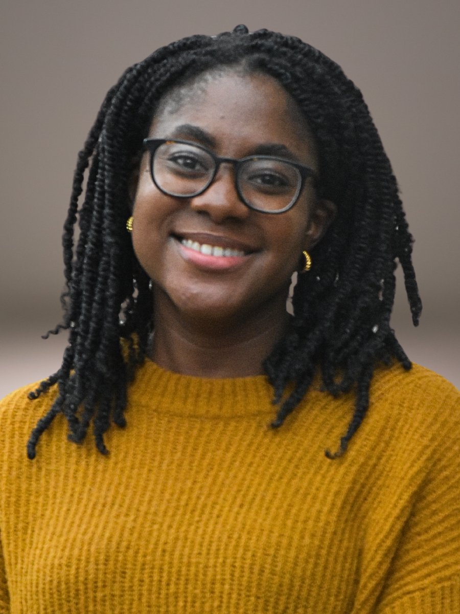 A smiling woman with dark skin, black glasses, black twisted hair, gold hoop earrings, and a mustard yellow sweater. She volunteers at Grace Capital City, a church in Washington, DC.