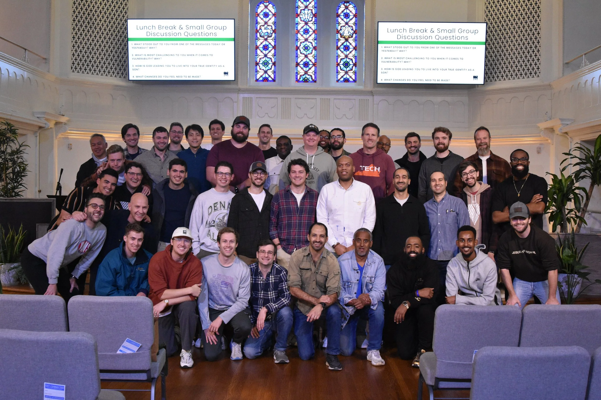 Group of diverse men gathered for a photo in a church with stained glass windows. Some are sitting, others standing, smiling for the camera. Taken after the Men's Conference for Grace Capital City, a church in Washington, DC.