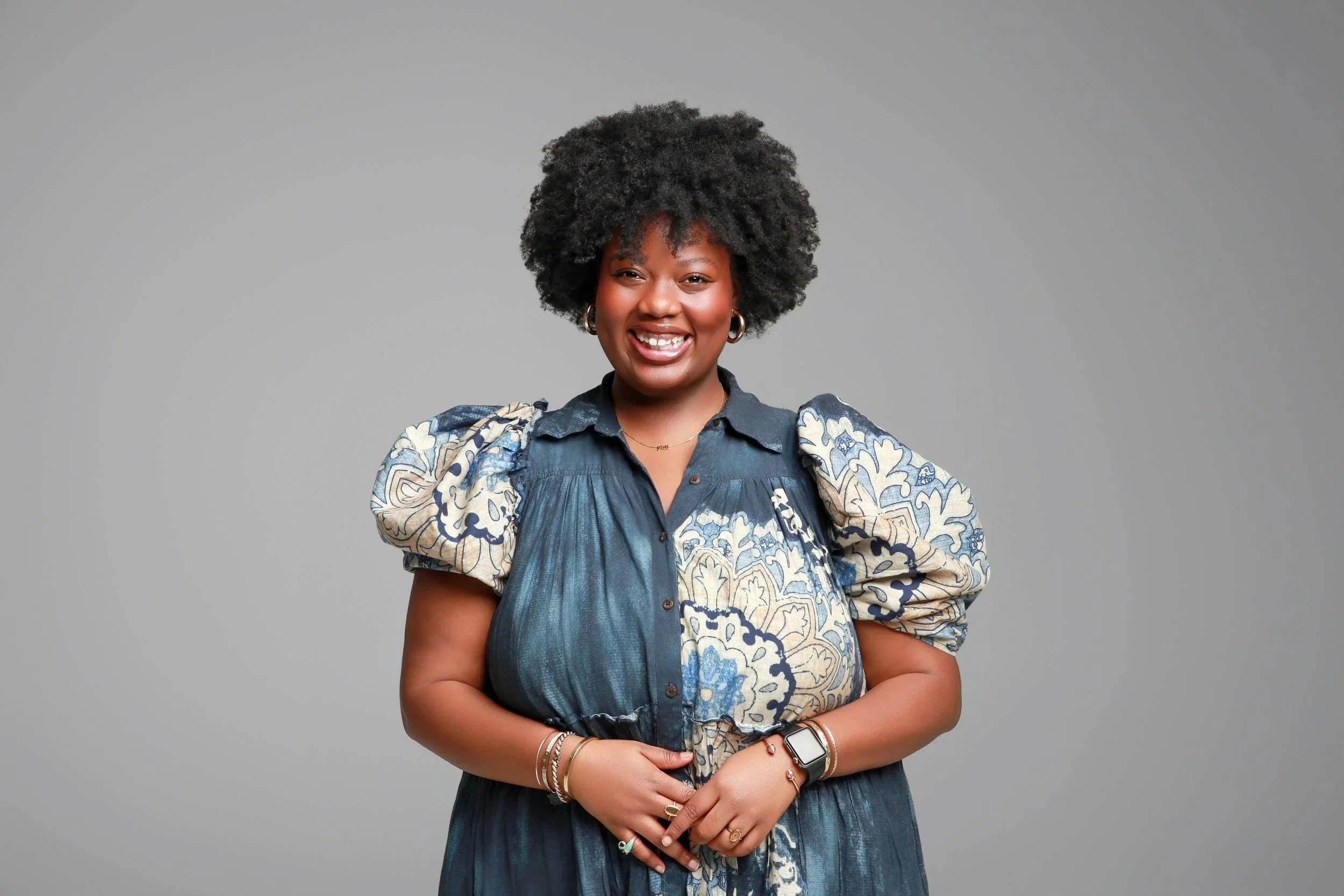 A smiling, black woman with curly black hair wearing a blue patterned dress standing against a gray background. She serves on staff at Grace Capital City, a church in Washington, DC.