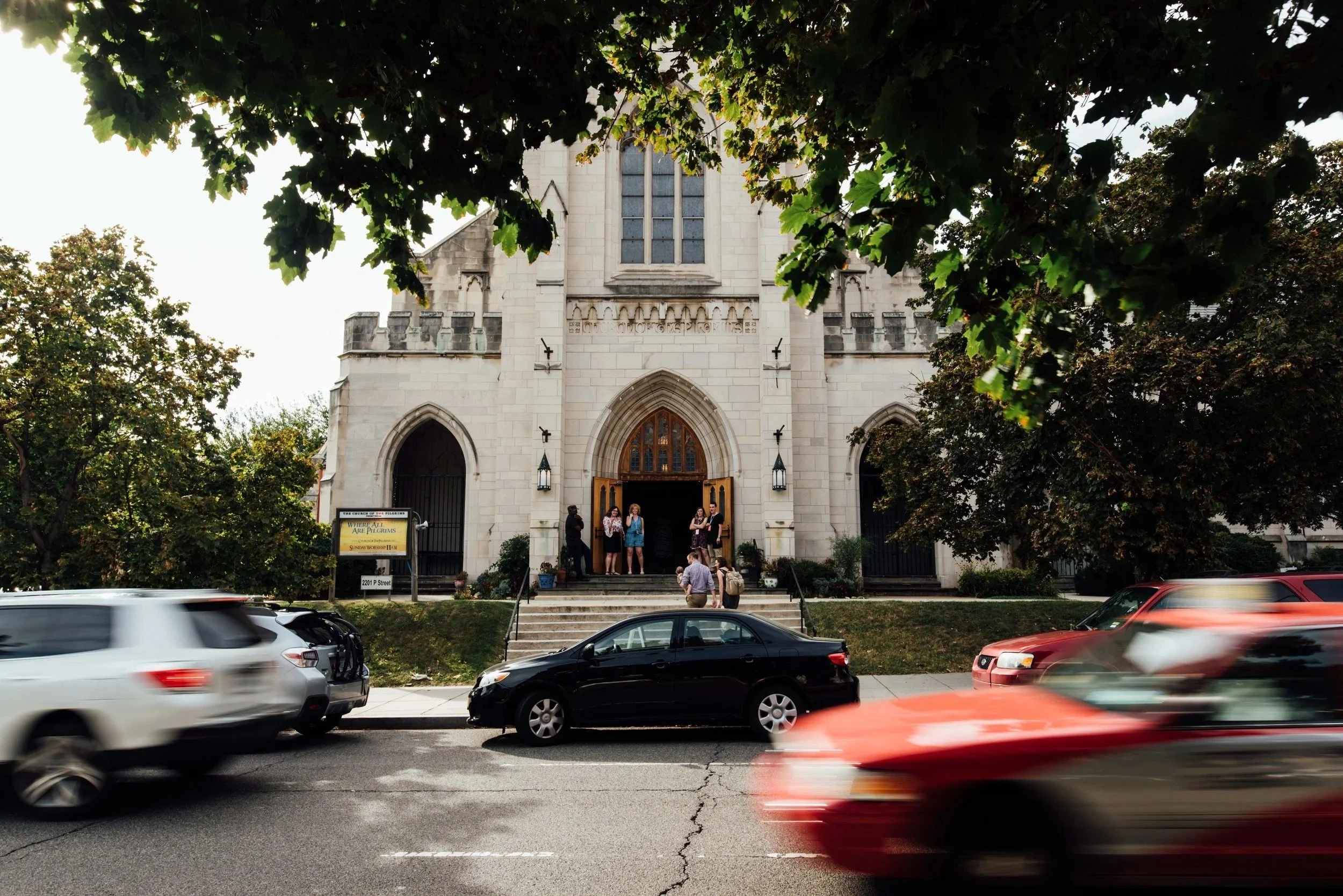 A church with gothic architecture style, light-colored stone exterior, large stained-glass window, and open wooden doors. Several people are standing on the steps, and cars are passing by on the street in front of a Grace Capital City church service.