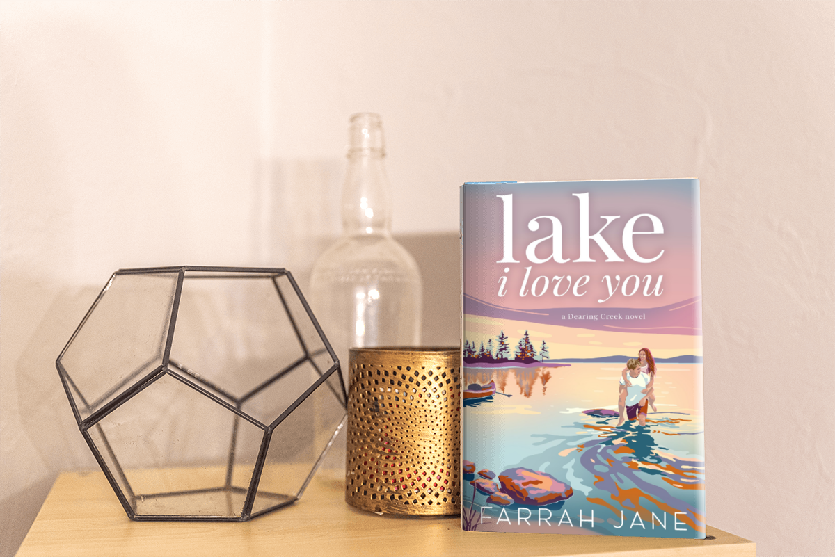 A paperback book titled 'Lake I Love You' by Farrah Jane standing upright on a wooden surface, with a decorative geometric metal object, a gold candle holder, and an empty glass bottle in the background.