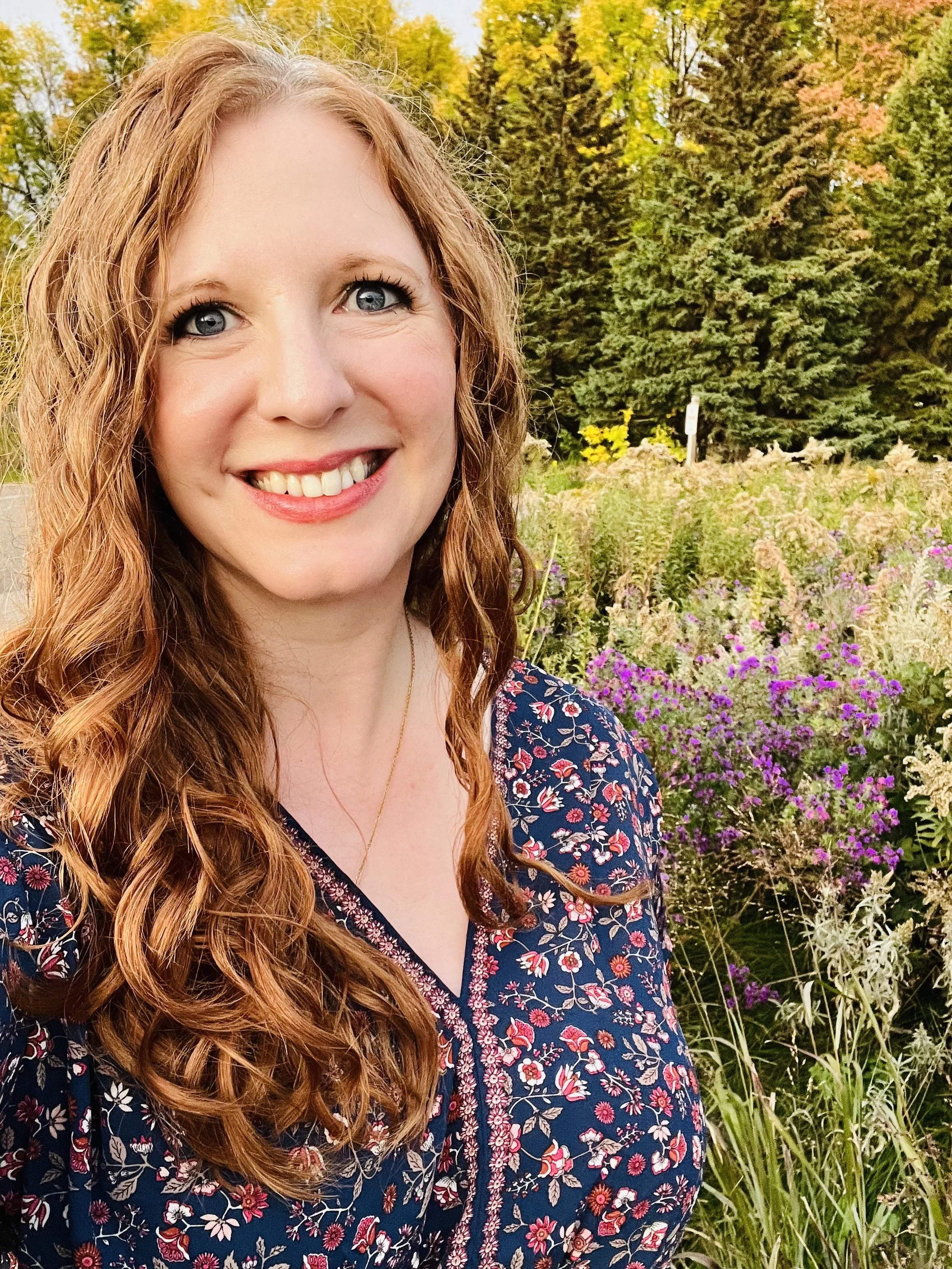 A smiling woman with curly red hair wearing a floral blouse standing outdoors in front of a garden with colorful flowers and trees.