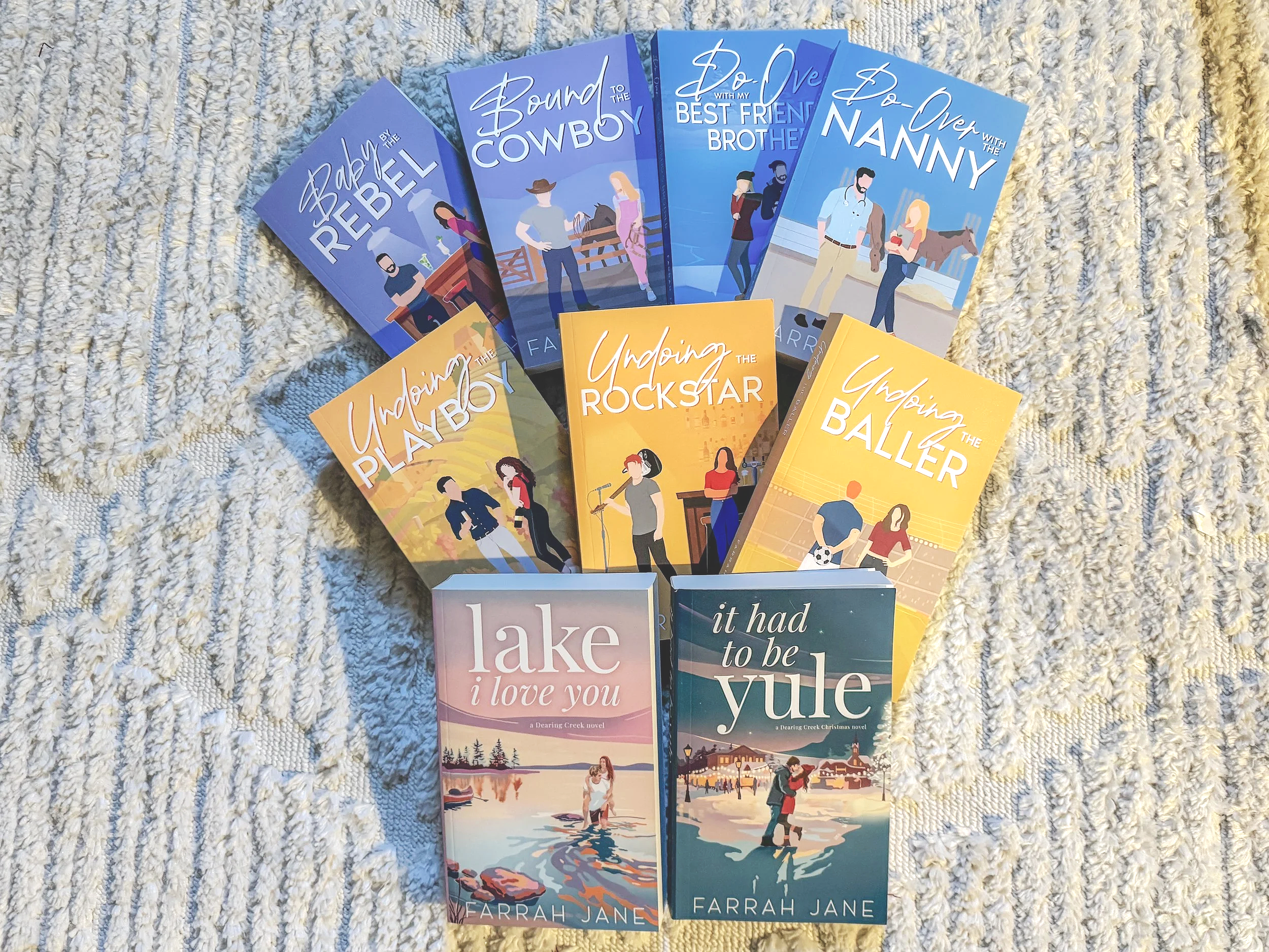 Collection of eleven colorful illustrated paperback books arranged on a textured white and beige surface, featuring titles like "Baby Rebel," "Bound to the Cowboy," "Do Over with My Best Friend Brother," "Do Over with the Nanny," "Undaing Playboy," "Undaing Rockstar," "Undaing the Baller," "Lake I Love You," and "It Had to Be Yule."