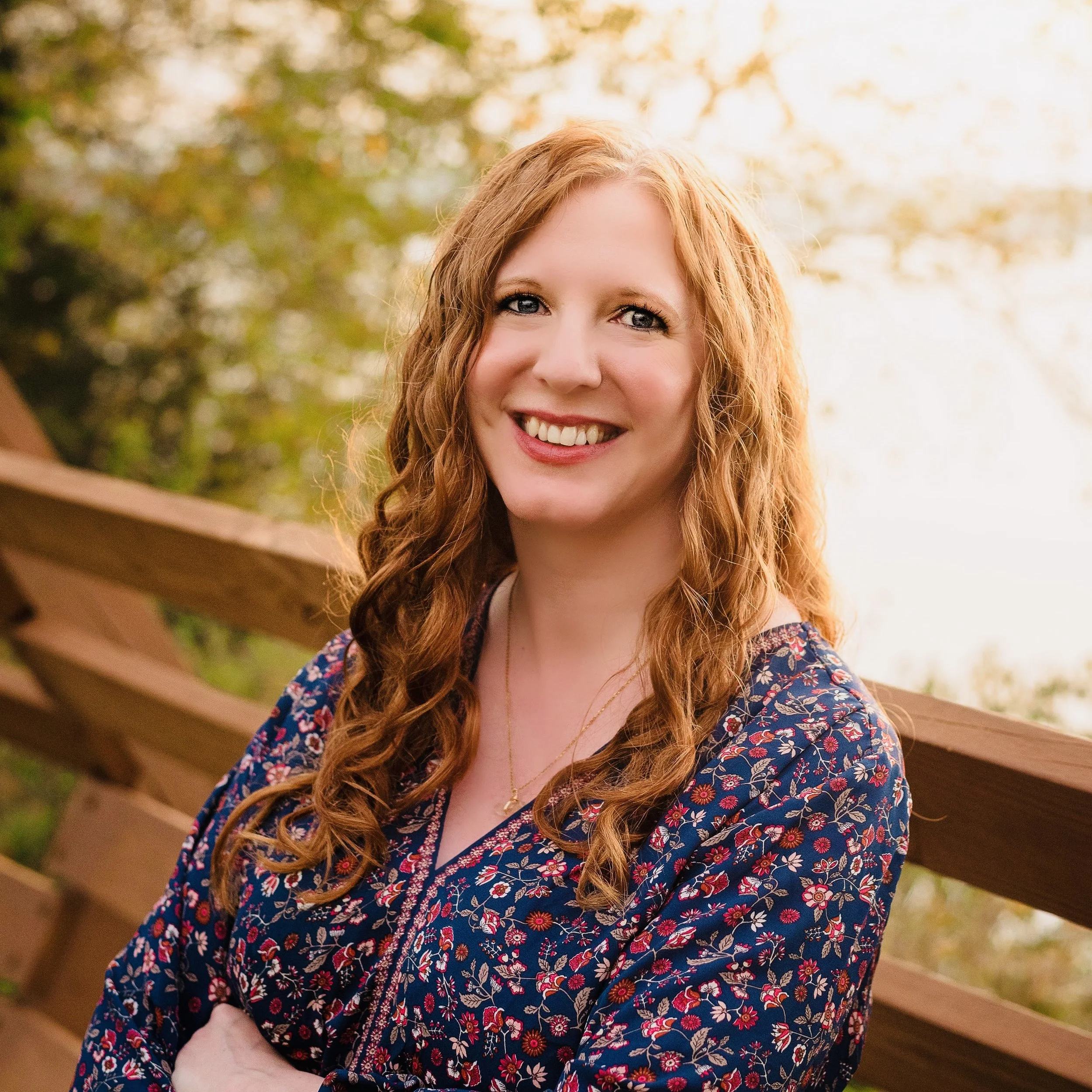 A woman with long, curly red hair smiling outdoors in front of a wooden railing and trees with fall foliage.