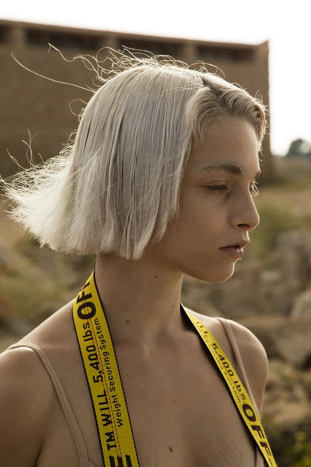 Ragazza con capelli biondi corti, vestita con una tuta beige e una cinghia gialla con scritte nere, in un ambiente naturale con sfondo sfocato.