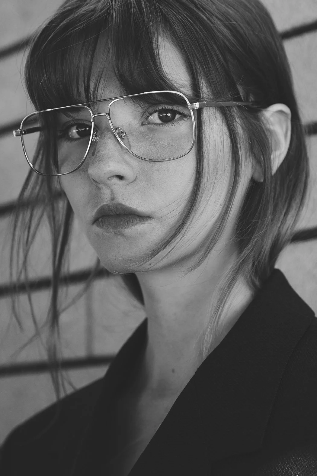 Ritratto in bianco e nero di una giovane donna con occhiali di metallo, capelli corti e sfumati, che guarda verso la fotocamera con un sorriso sottile, sfondo di una parete di mattoni.