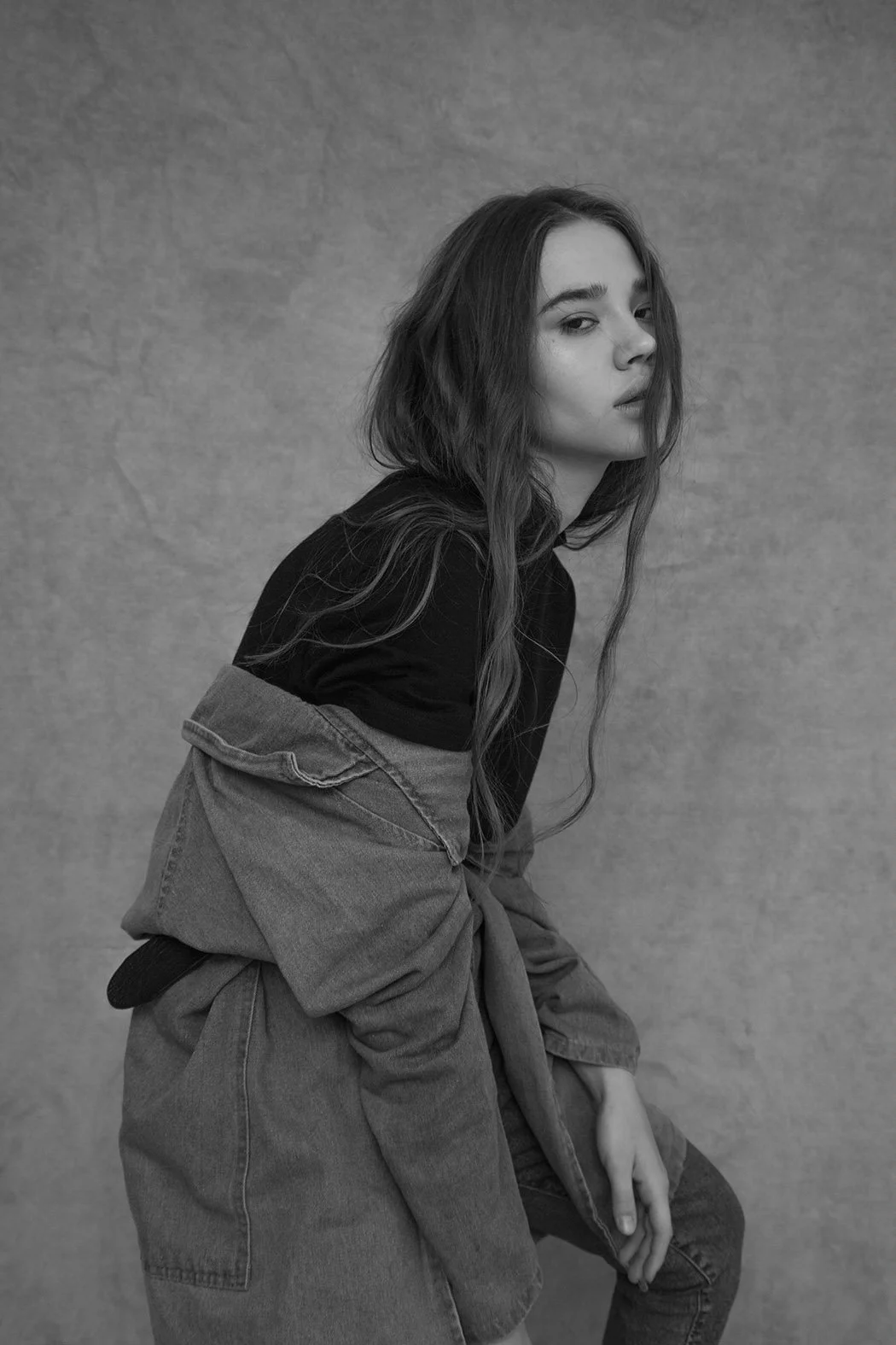 Ragazza con capelli lunghi e castani che indossa una giacca di jeans sbottonata e una maglietta nera, posa contro una parete liscia di colore chiaro, in bianco e nero.