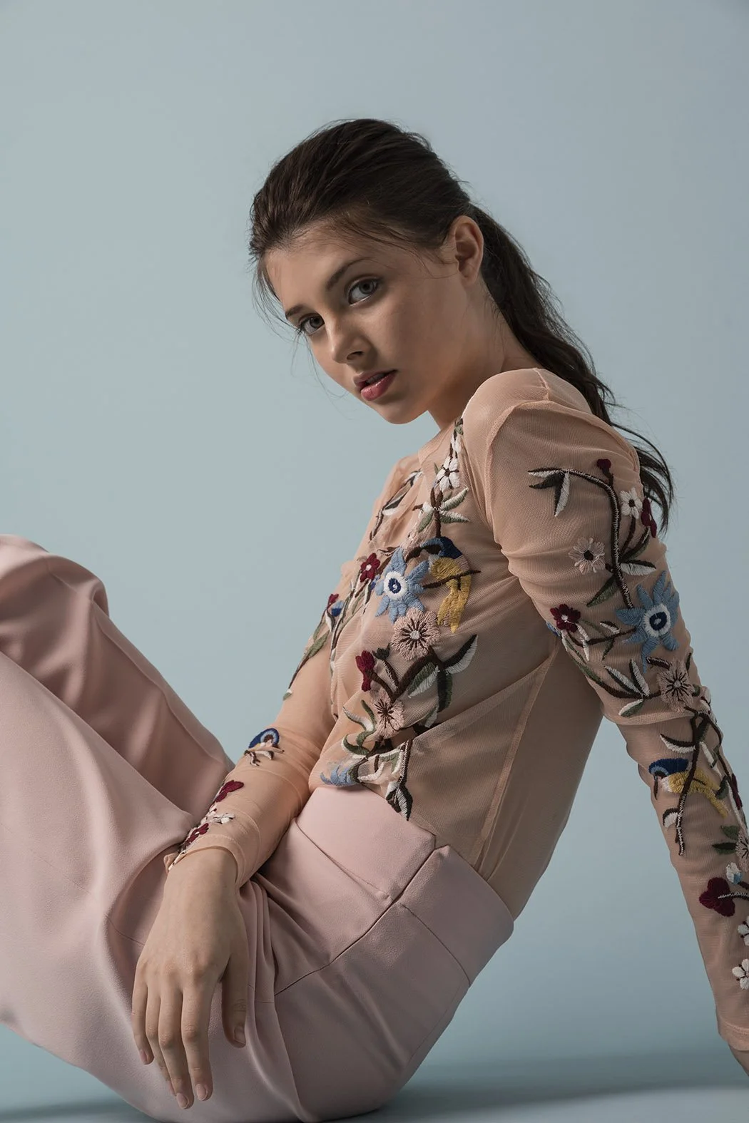 Ragazza seduta su sfondo azzurro, indossa un top rosa con ricami floreali e pantaloni rosa chiaro, guarda verso la fotocamera.