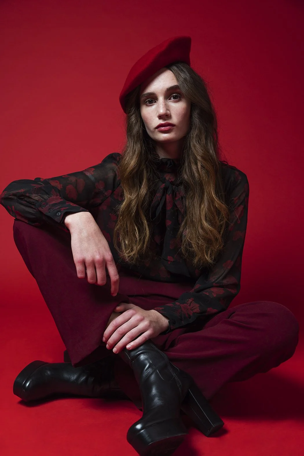 Una donna seduta sul pavimento con uno sfondo rosso, indossa un cappello rosso, una camicetta nera con motivi rossi e pantaloni bavori, con stivali neri.