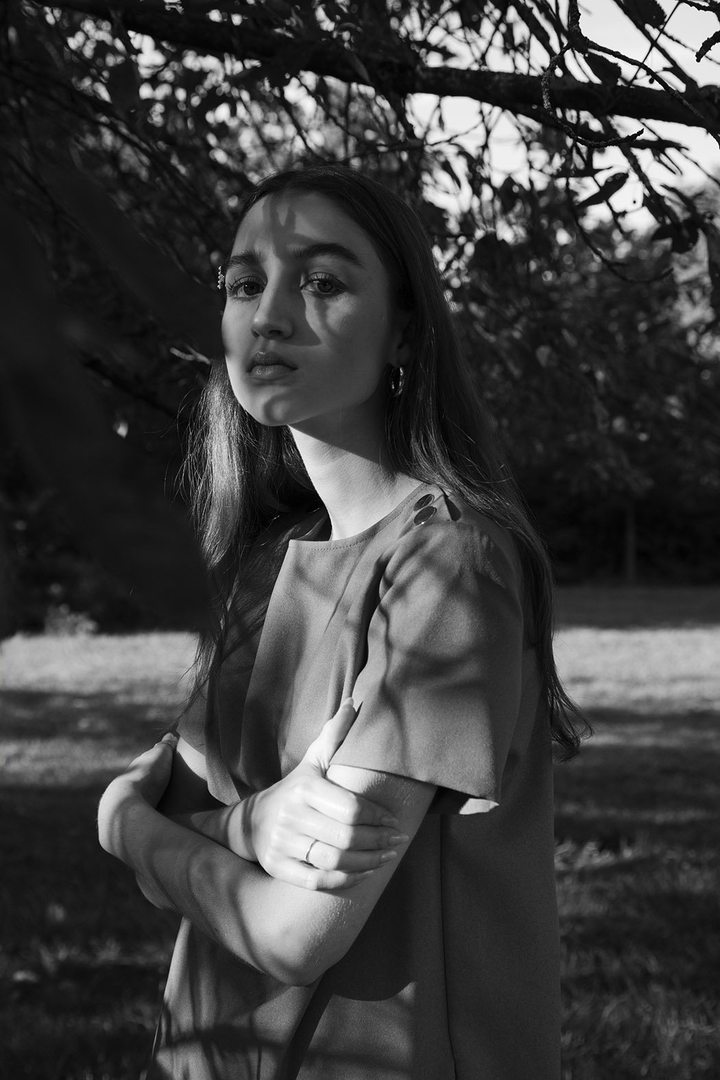 Ritratto di una donna con capelli lunghi, in posa in un'area all'aperto, con giochi di luce e ombre create dalle foglie di un albero.