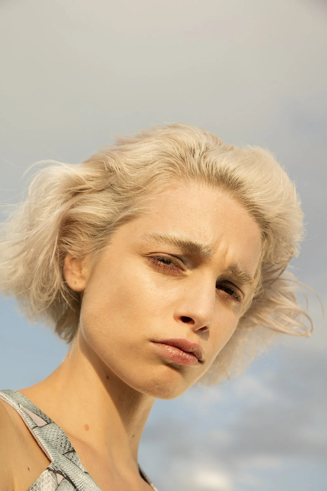 Ragazza dai capelli biondi corti con espressione seria o pensierosa contro uno sfondo di cielo