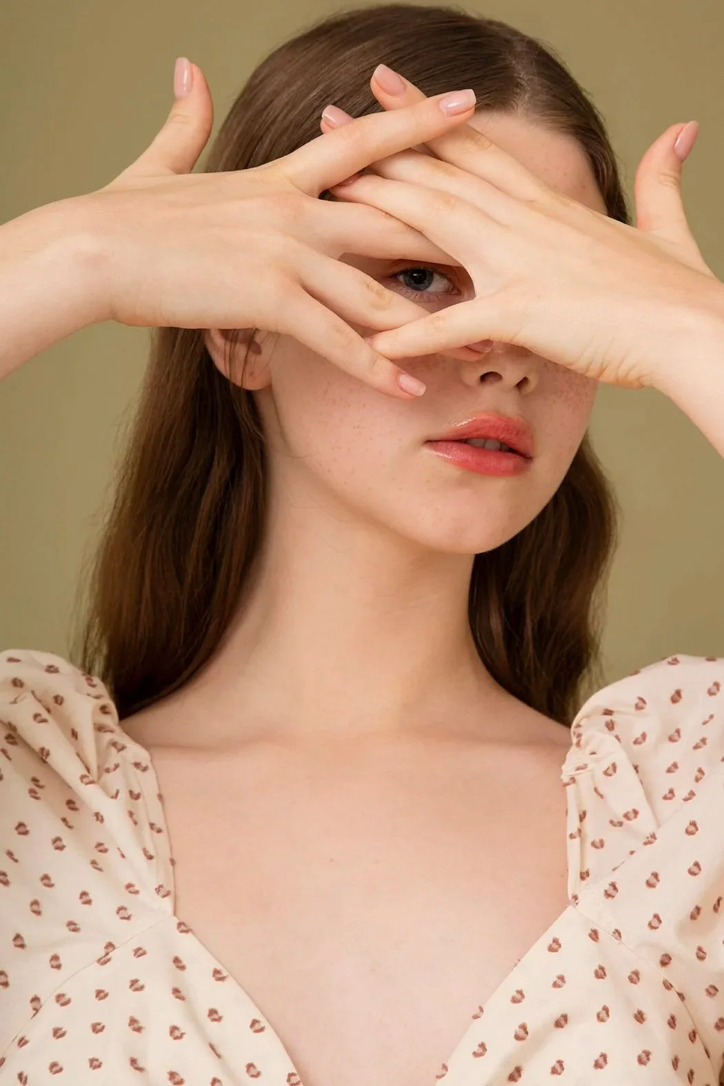 Ragazza con capelli castani, che indossa una maglietta crema con motivo a pois rossi, si copre gli occhi con le mani e guarda verso l'obiettivo.