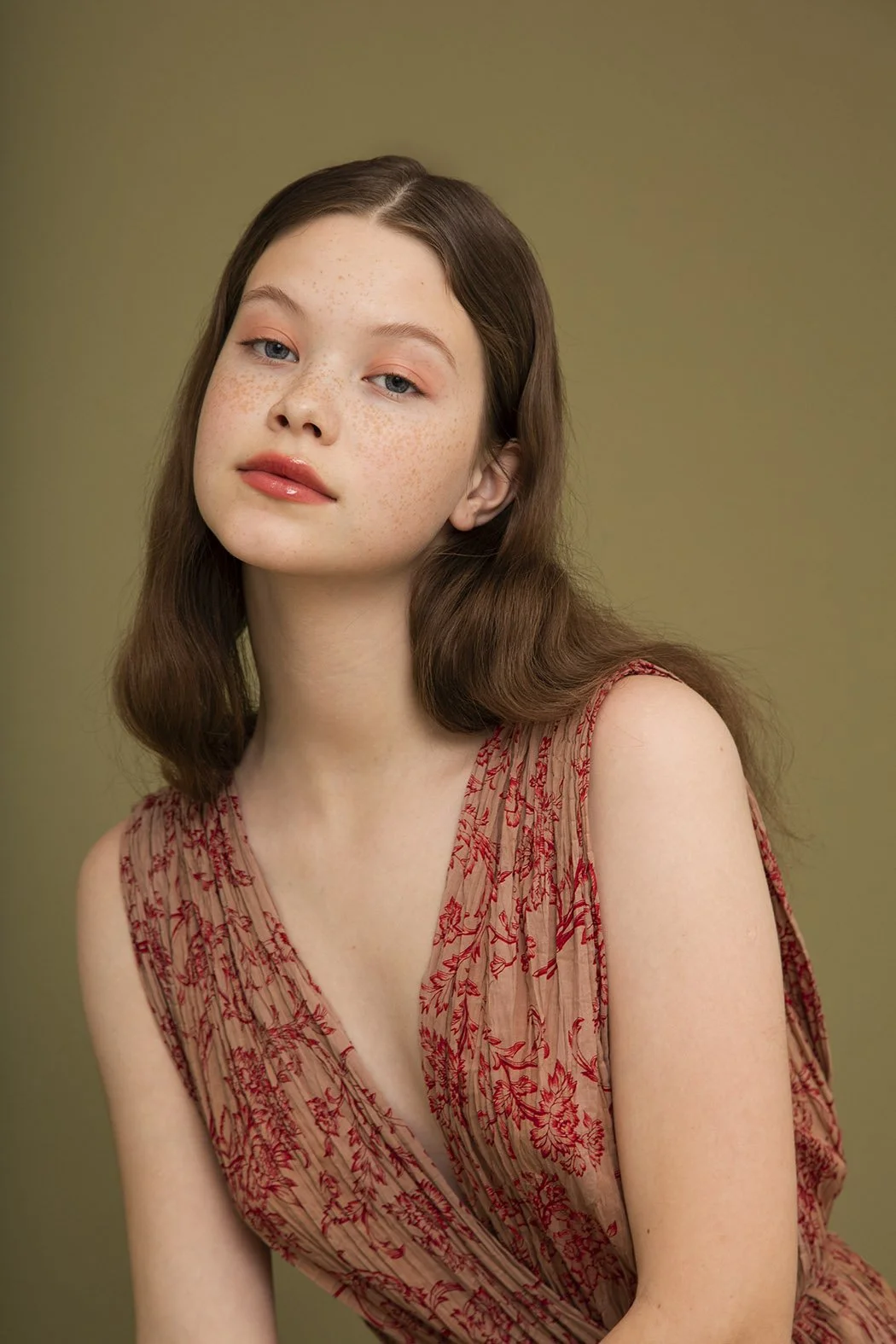 Ragazza con capelli castani, pelle chiarissima e lentiggini, indossa un vestito senza maniche con fiori rossi, sfondo verde oliva.