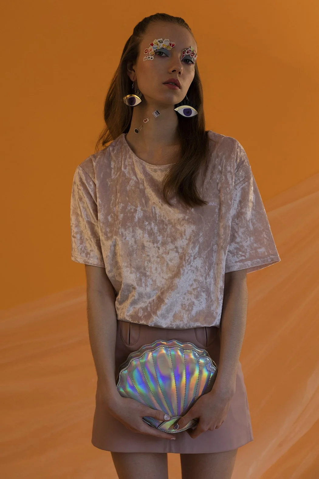 Una donna con abbigliamento rosa, orecchini a forma di occhi e una borsetta a forma di conchiglia iridescente, su uno sfondo arancione.