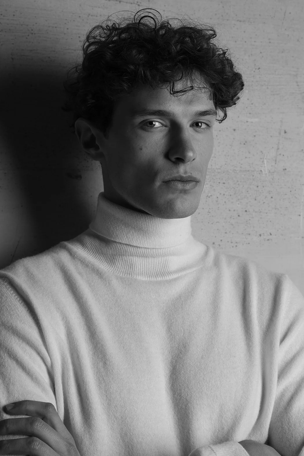 Ritratto di un giovane uomo con capelli ricci, vestito con una maglia a collo alto, sfondo chiaro e lucido.
