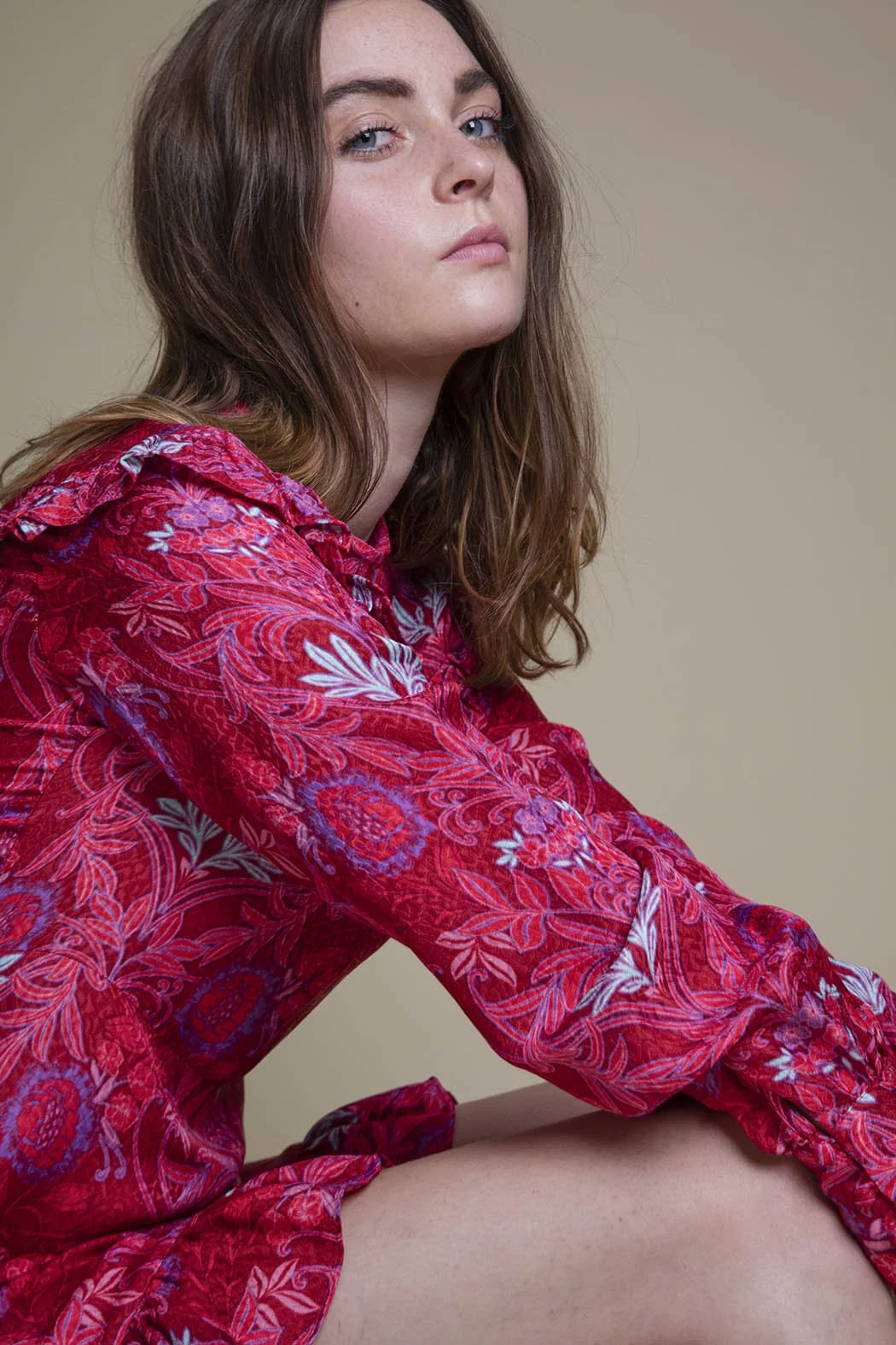 Ragazza con i capelli castani e ricci, vestita con un abito rosso a fiori, che guarda verso l'obiettivo in un ambiente neutro.
