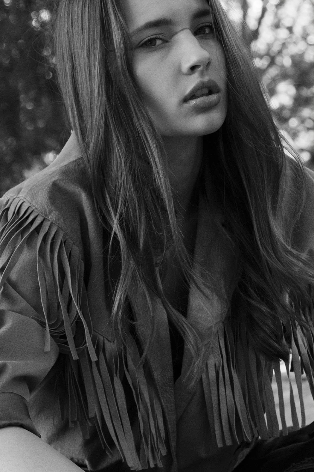 Ragazza con capelli lunghi e mossi, occhi acuti, abbigliamento con frange sulle spalle, in posa in un ambiente naturale.