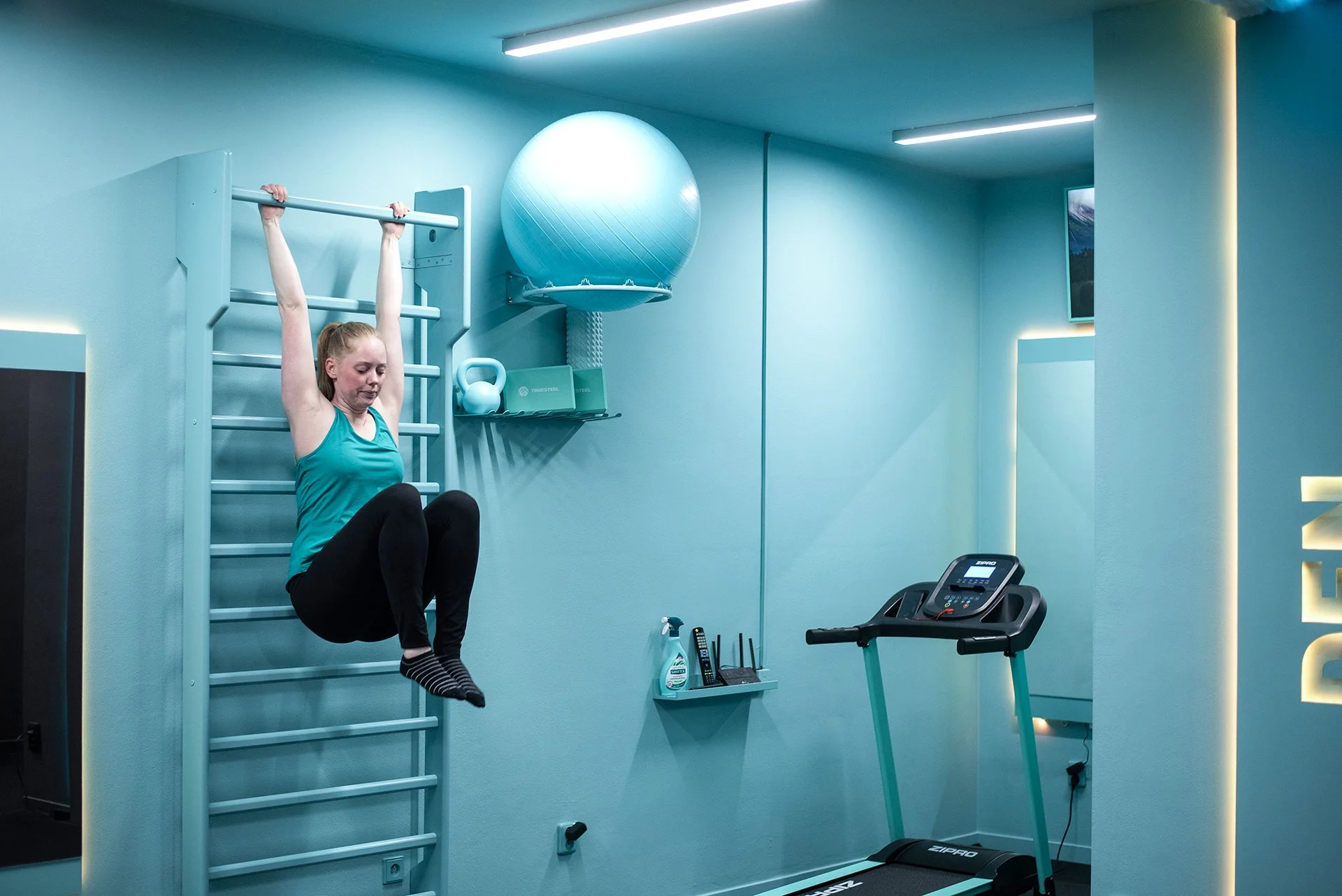 A woman exercising on a gym wall ladder, hanging from a pull-up bar, with a large blue exercise ball and gym equipment on the wall behind her in a turquoise-colored room.