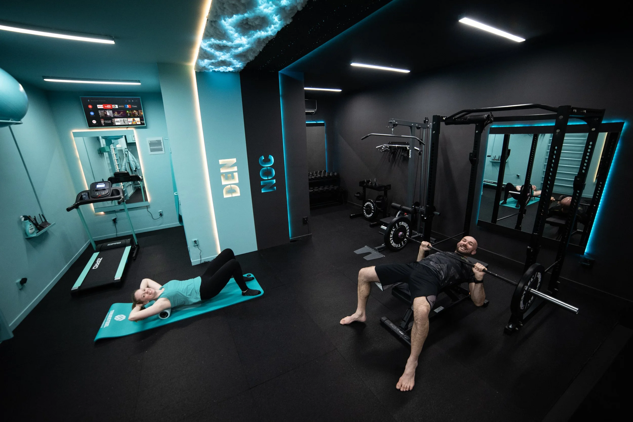 A modern gym with black and teal walls, featuring a treadmill and weightlifting equipment. Two people are exercising: a woman lying on a teal yoga mat doing a crunch with a foam roller beneath her lower back, and a man lying on a padded bench holding a barbell across his shoulders, smiling. The gym has a reflective mirror, LED lighting, and a ceiling decorated with clouds and star-like lights.