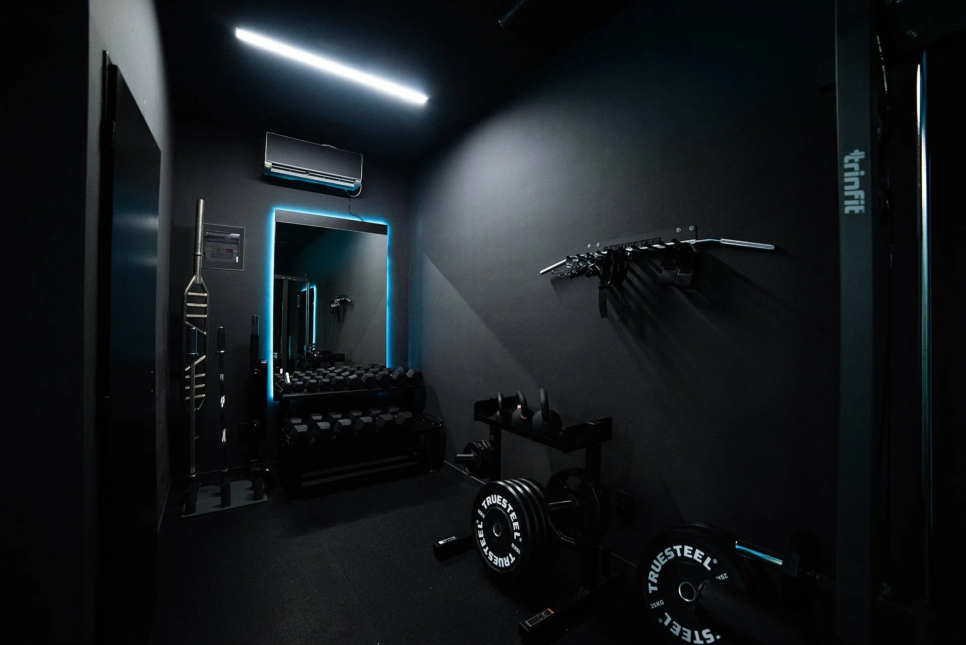 A dark modern home gym with a mirror illuminated by blue LED lights, dumbbells, kettlebells, and weight plates, with fitness equipment neatly arranged.