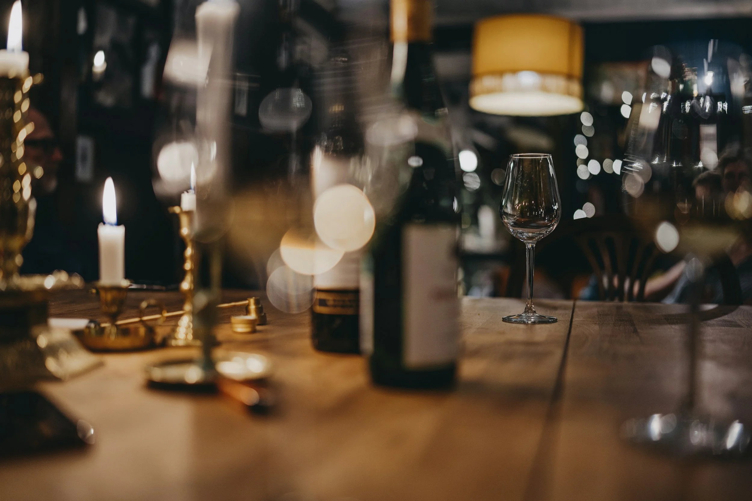 A dimly lit restaurant or bar with a wine glass on a wooden table, surrounded by blurred candles, bottles, and people in the background, creating a cozy ambiance.