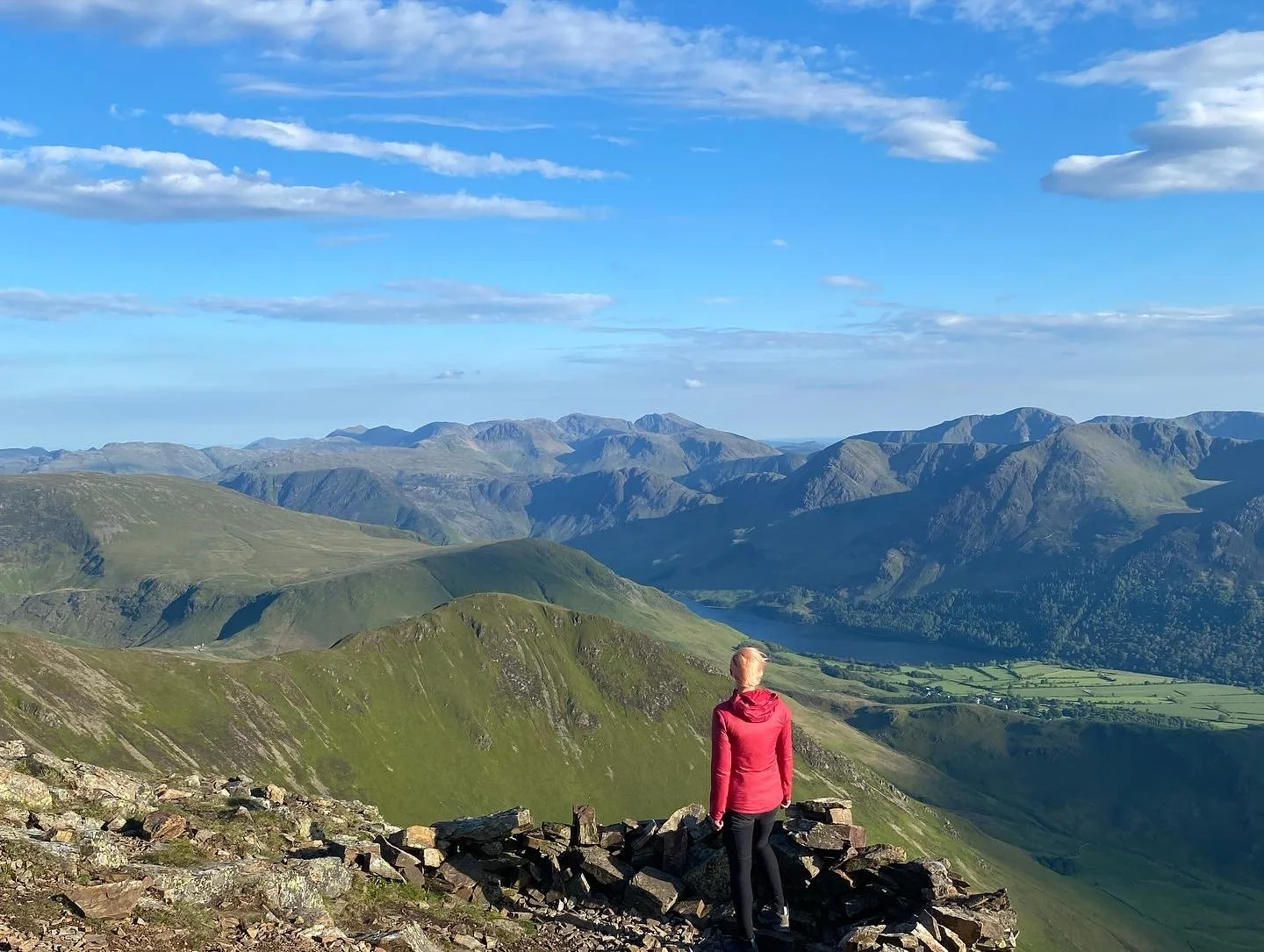 Person in a pink jacket standing on rocks overlooking green mountains and a blue lake under a partly cloudy sky.