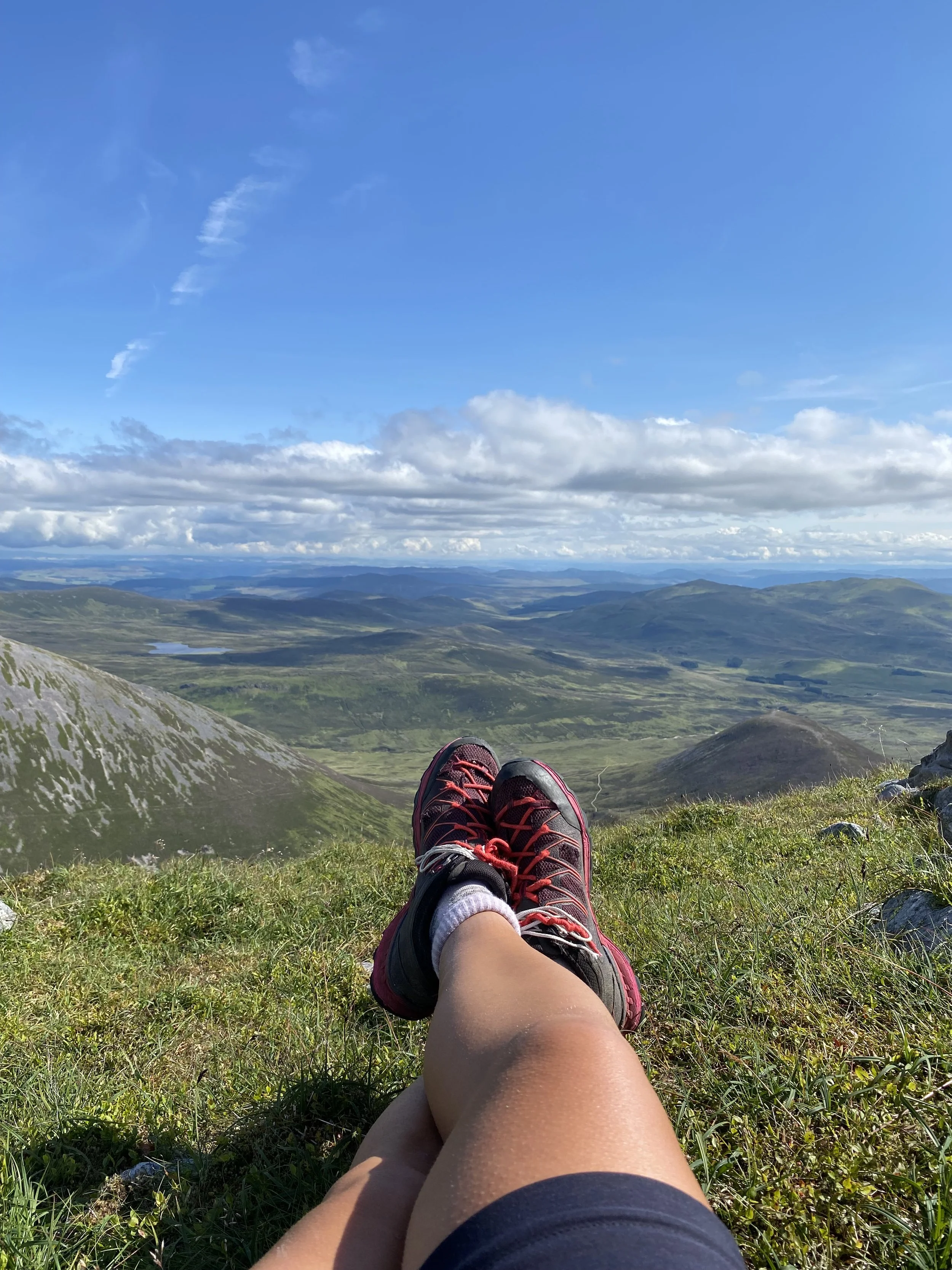 Person relaxing on a grassy hilltop overlooking a vast valley with rolling hills, a lake, and distant mountains under a partly cloudy blue sky.