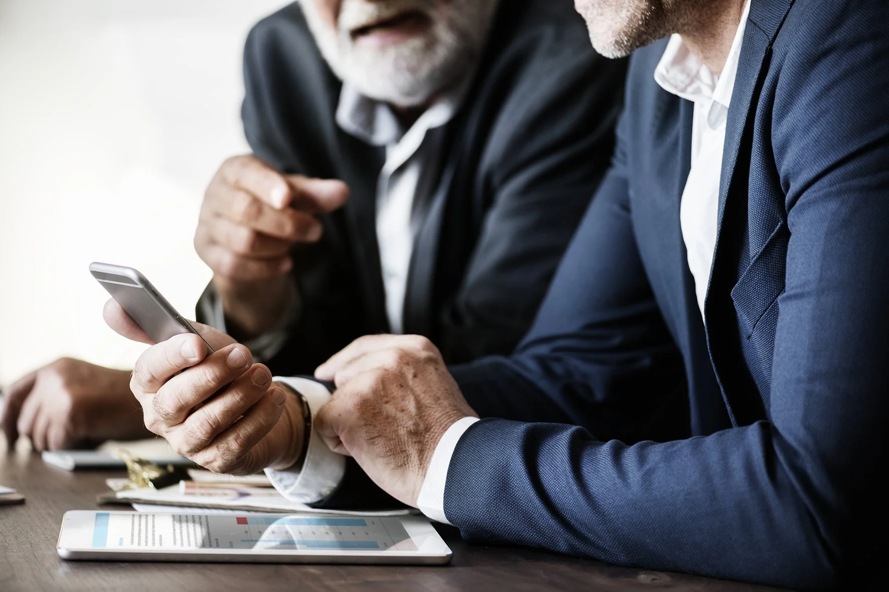 Two men in formal suits sitting at a table, looking at a smartphone together. There are documents and a tablet with charts on the table.