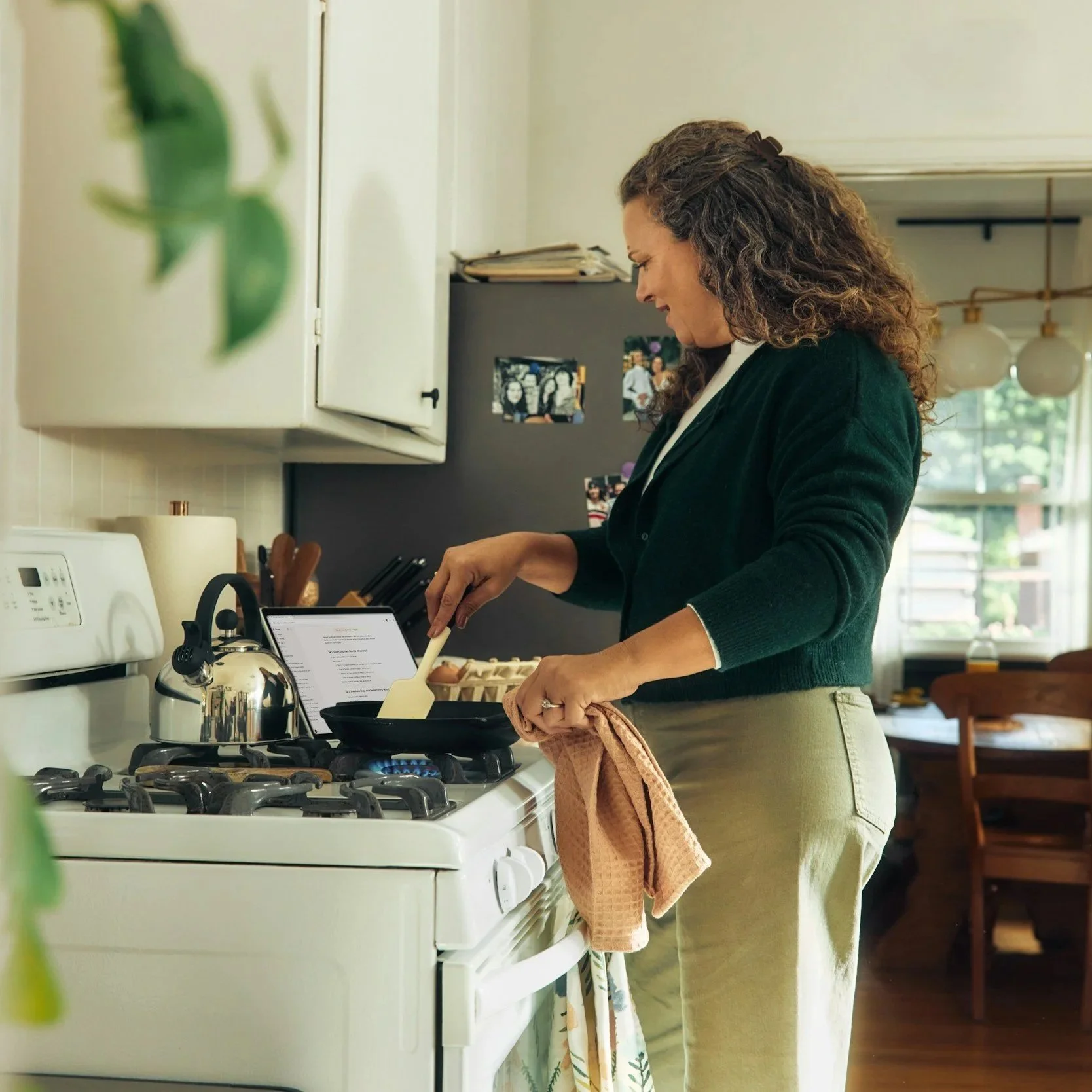 Woman cooking on stove in kitchen, holding a pink kitchen towel for drying. The woman has curly hair and is wearing a dark sweater and beige pants.