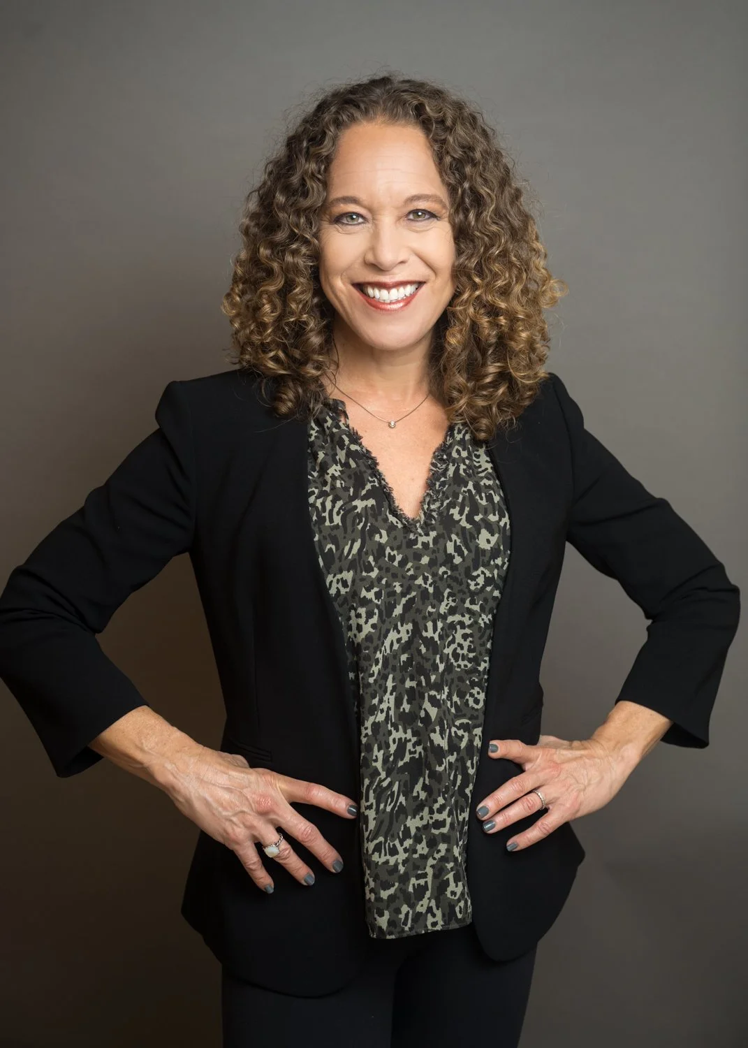 A woman with curly hair smiling, wearing a black blazer over a leopard print blouse, standing against a plain grey background.