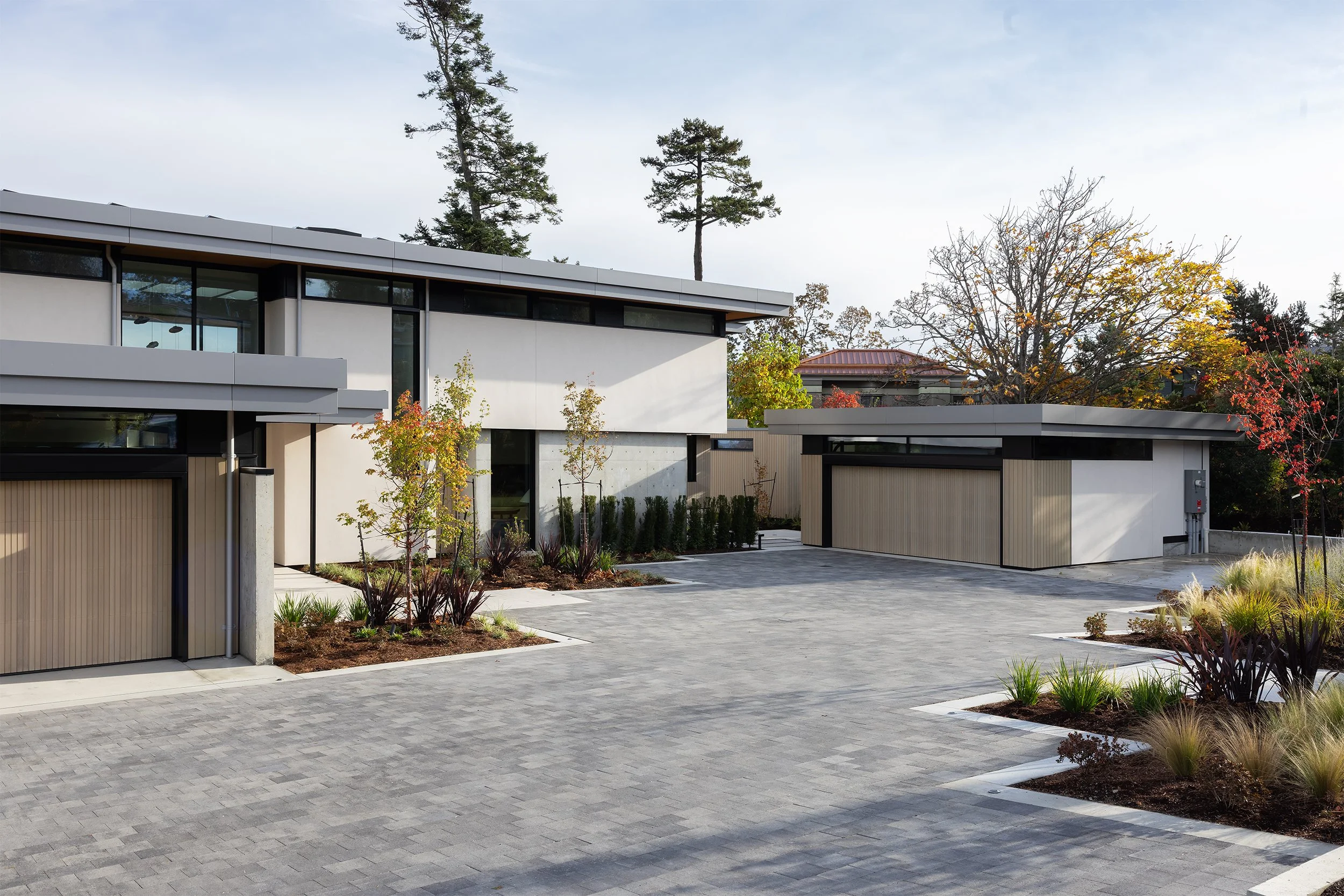 Modern residential house with flat roof, large windows, and landscaped yard with small trees and shrubs, surrounded by tall trees.