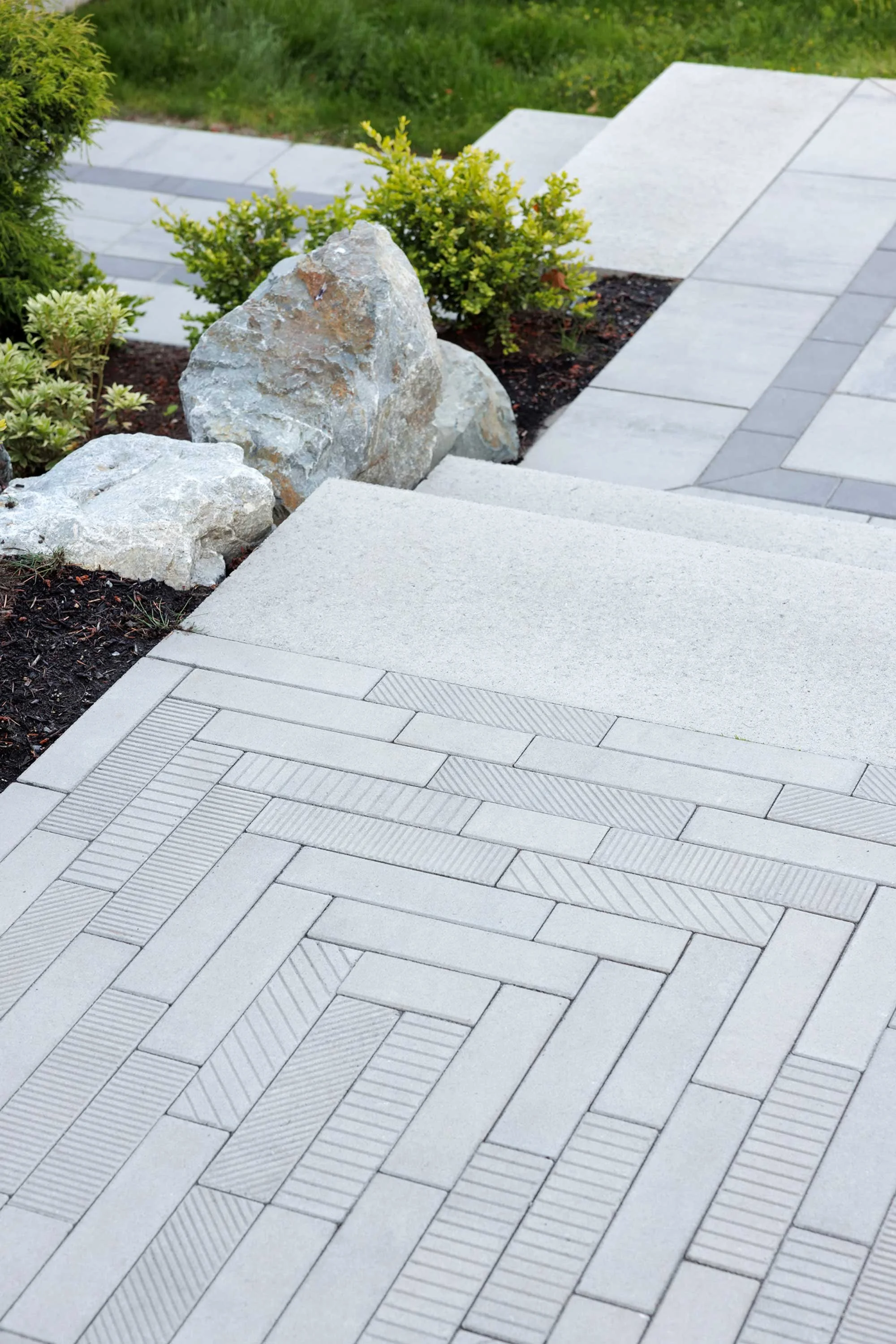 A paved pathway with gray bricks, leading to steps, surrounded by small green bushes and rocks, in a garden area.