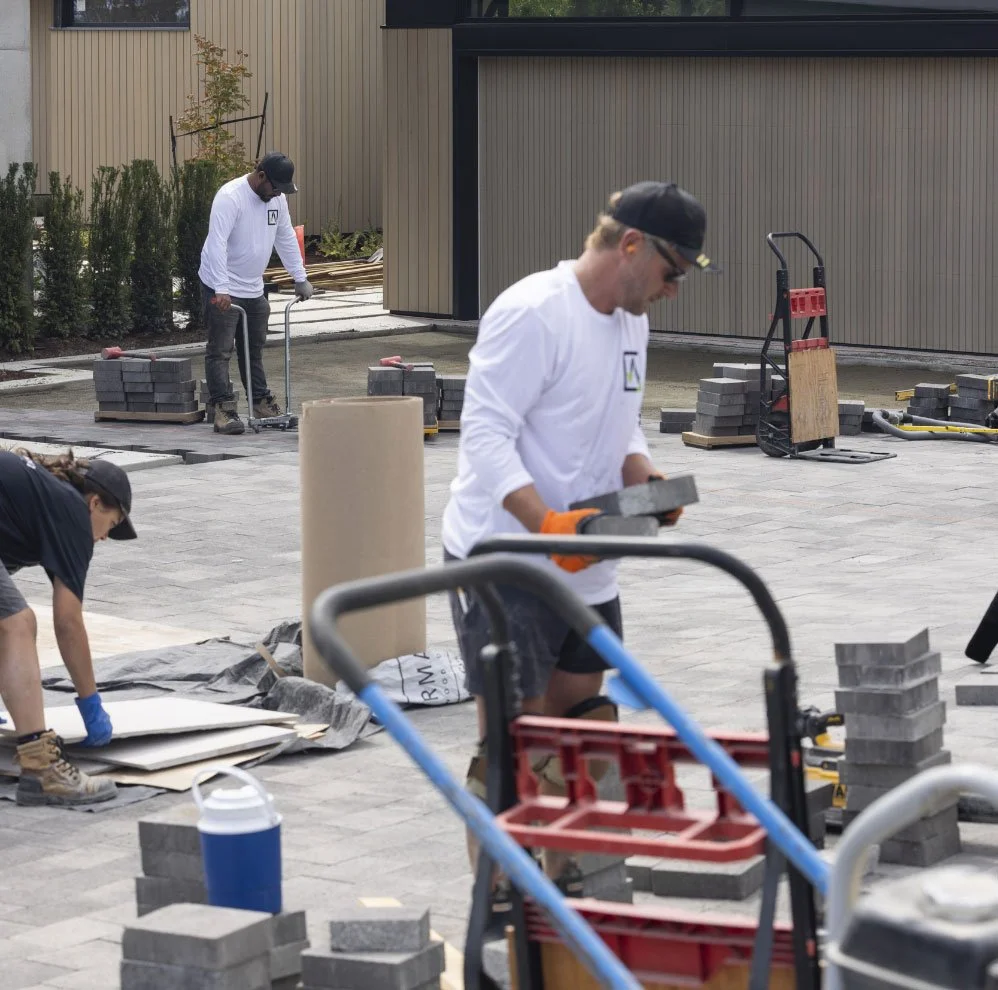 Alliston Stoneworks team laying paving stones outdoors, using tools and materials such as bricks, pallets, and wheelbarrows.