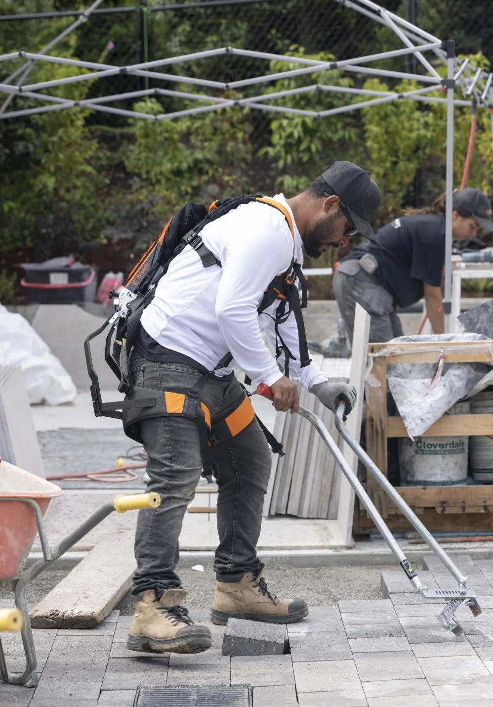 Man operating a manual brick splitter on a paved surface, working on hardscaping with greenery and other specialists nearby.