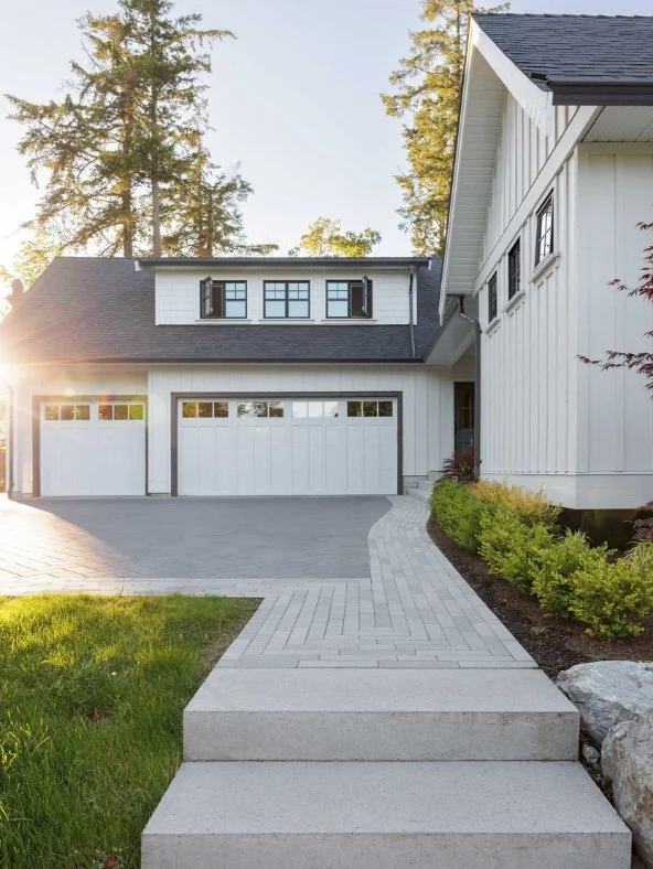 Front view of a modern white house with a garage, a brick-paved driveway, a concrete walkway leading to the entrance, and landscaped bushes, with tall trees in the background.