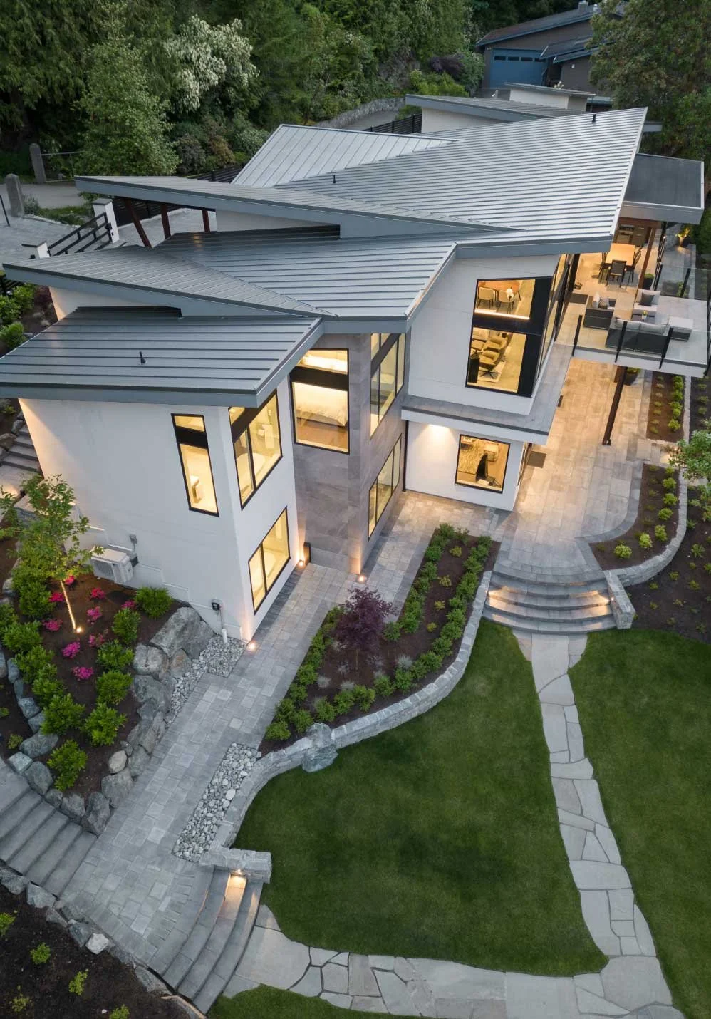 A modern multi-story house with large windows, and an outdoor patio area, surrounded by hardscaped gardens and walkways.