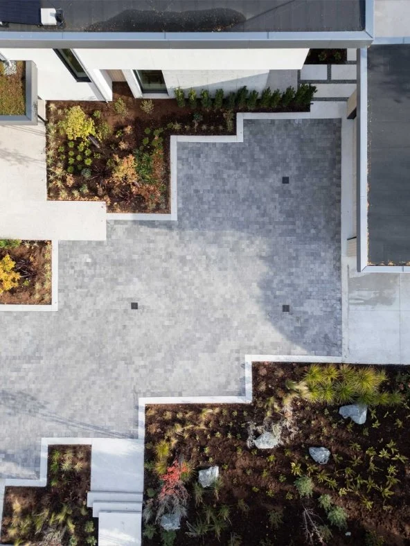 Overhead view of a modern residential courtyard with a paved patio, surrounding garden beds with plants and rocks, and part of a building with large windows.