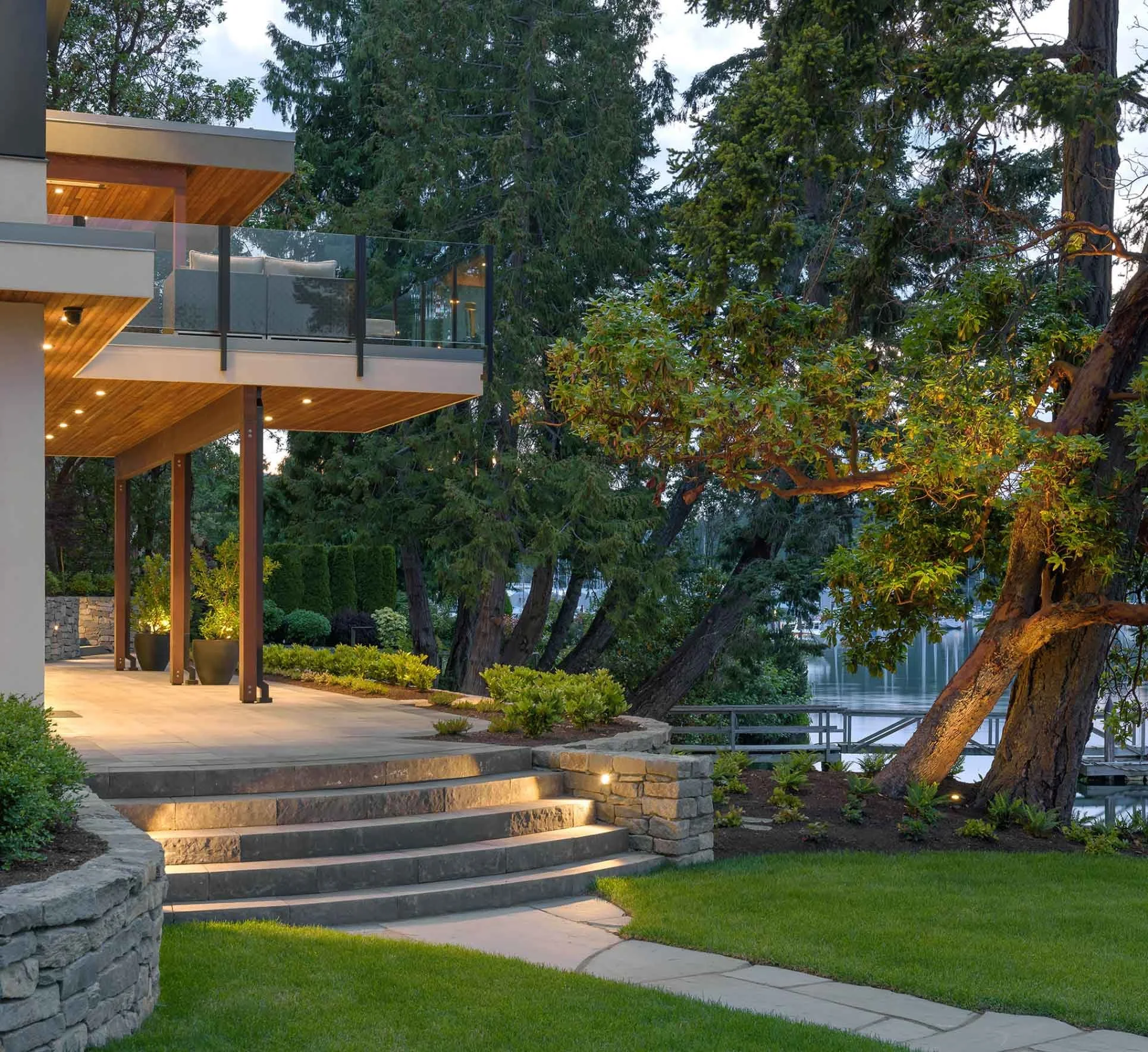 Modern house with large glass balcony, and outdoor lighting surrounded by trees and greenery by the ocean at dusk.