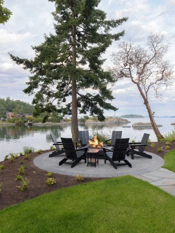 Waterfront backyard with a fire pit surrounded by eight black chairs, tall pine and leafless trees, green lawn, rocks, and a cloudy sky.