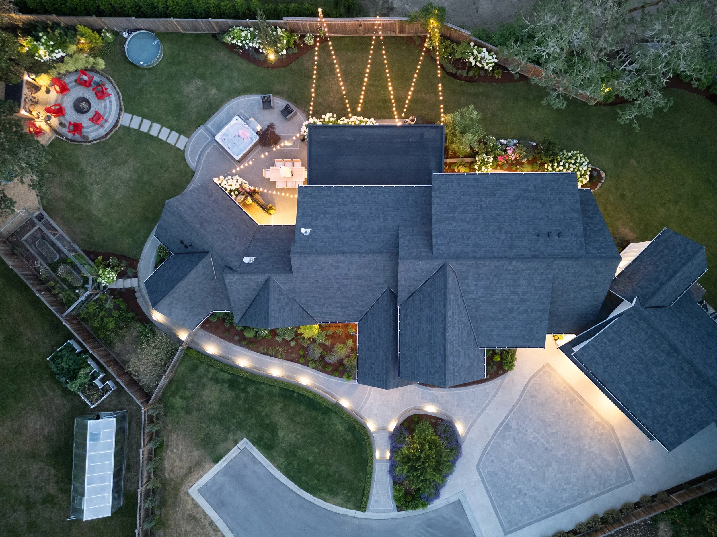 Aerial view of a backyard at dusk showing a lawn, garden beds, a patio with string lights, a hot tub, a fire pit with chairs, and paved walkways surrounded by trees and a wooden fence.