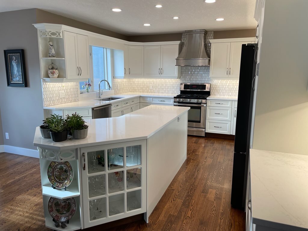 Modern white kitchen with island, wooden floors, stainless steel appliances, and decorative shelves.
