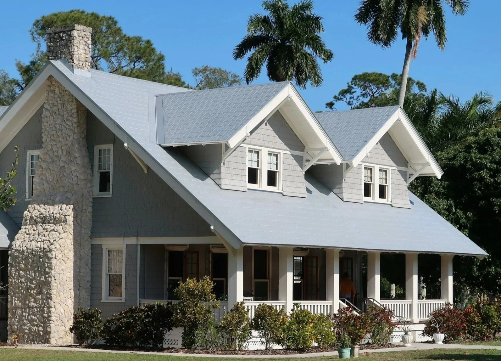 A large two-story house with gray exterior siding, a blue metal roof, and a covered front porch with white railings, surrounded by bushes and palm trees under a clear blue sky.