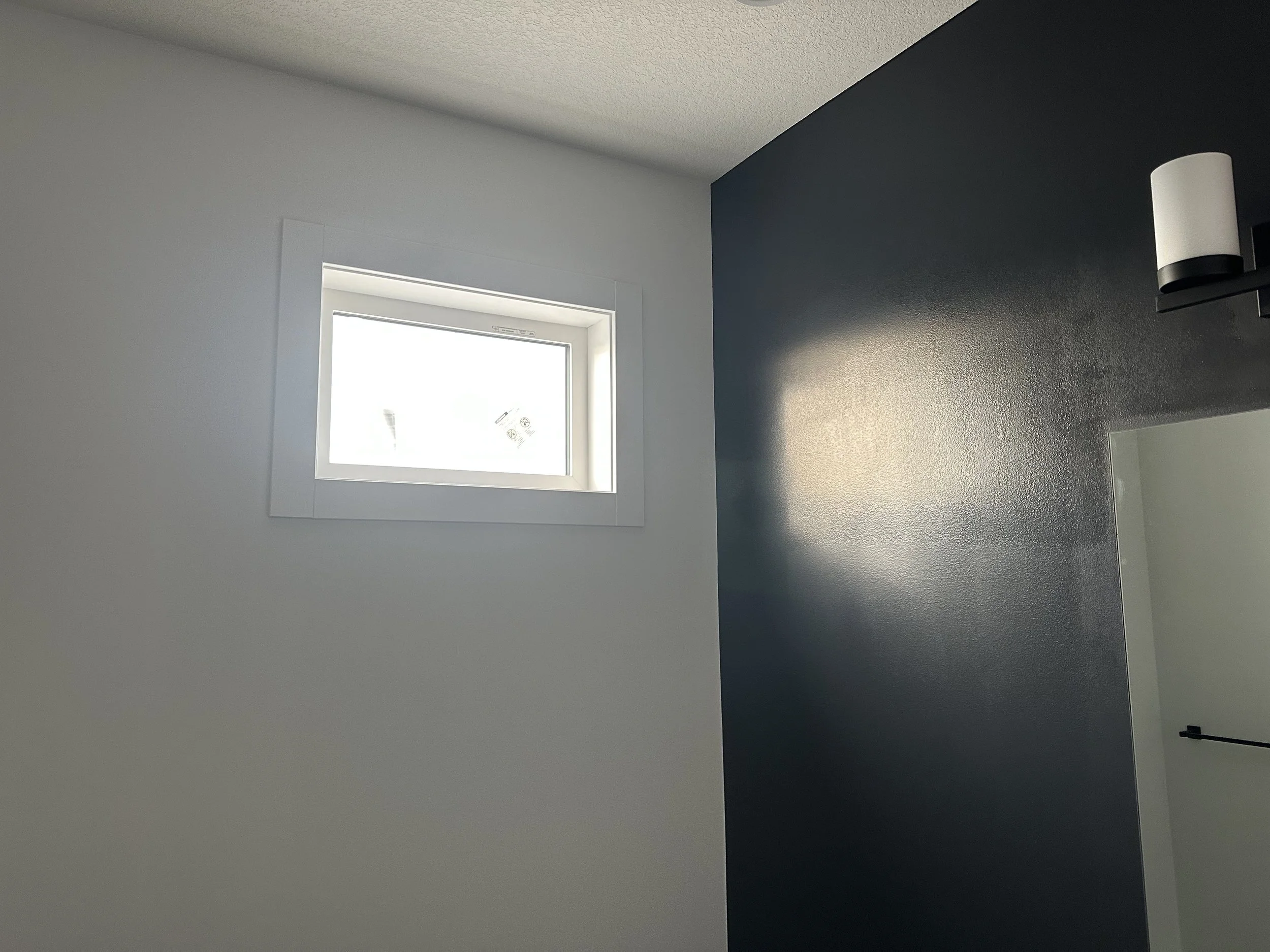 Interior corner of a room with a small window, white wall on the left, and dark gray wall on the right, with a wall-mounted light fixture on the right wall.