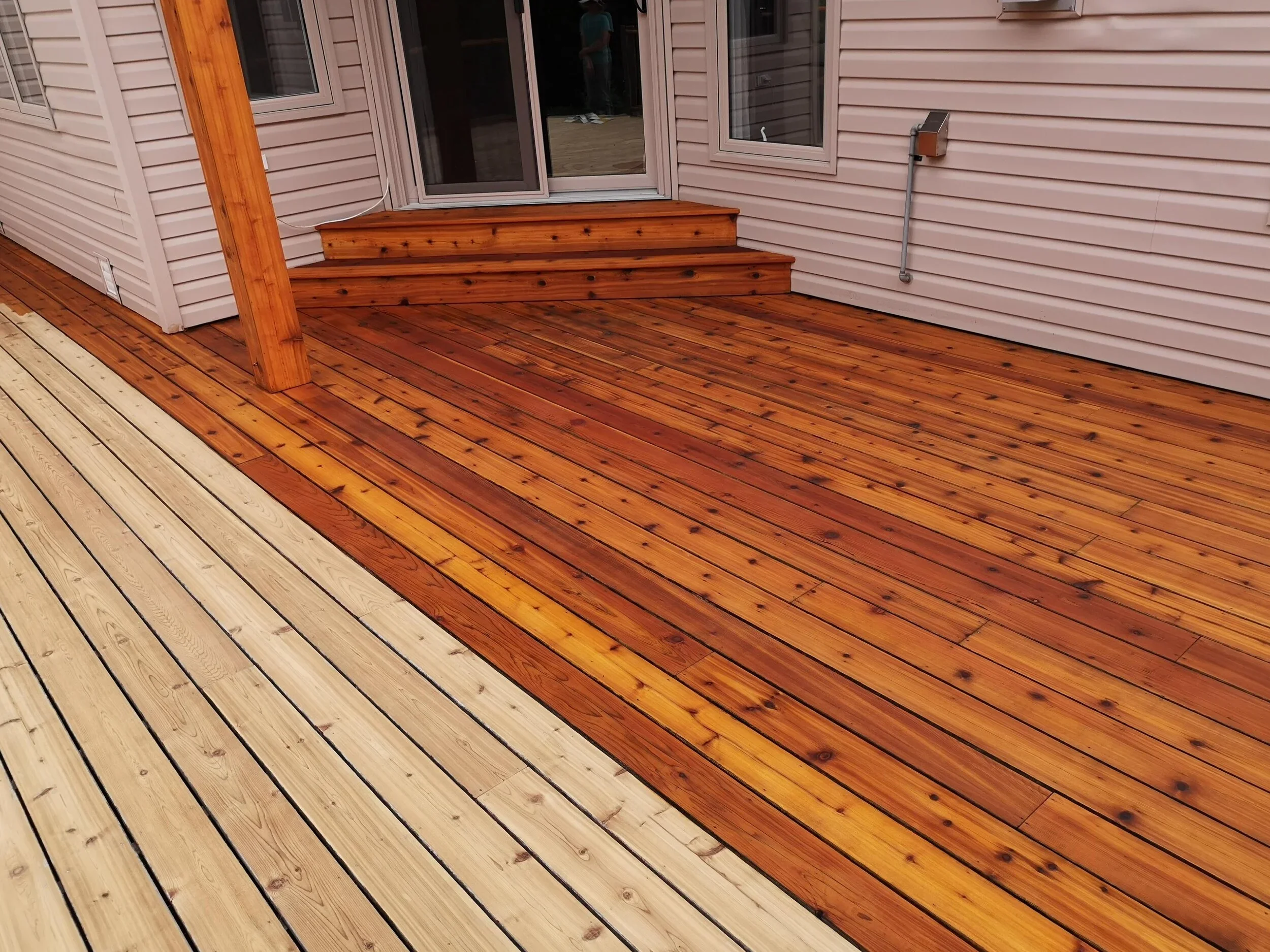Newly installed wooden deck with a mix of light and dark stained planks adjacent to a house with beige siding and sliding glass door containing three steps.
