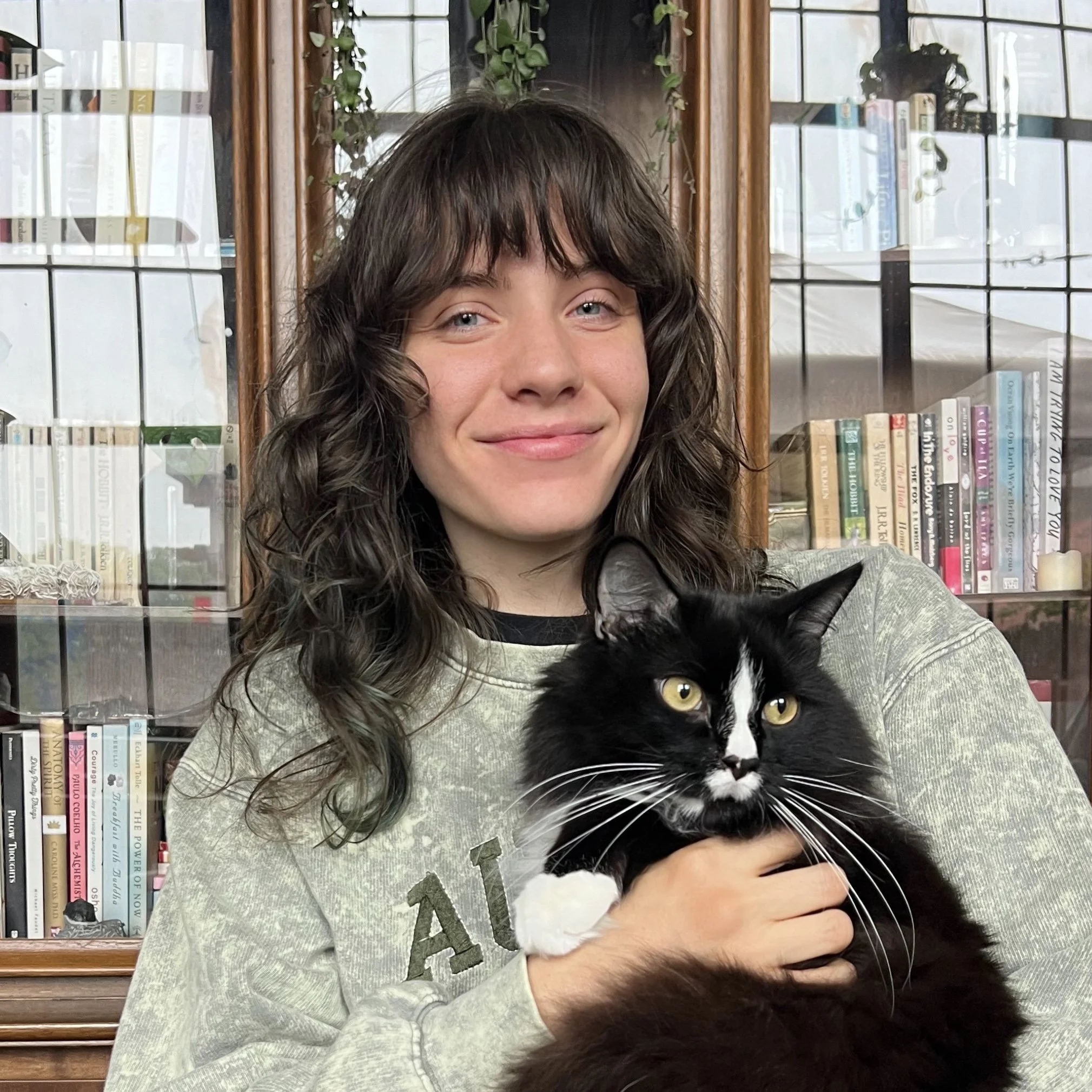 A young woman with wavy brown hair and a light gray sweatshirt holding a black and white cat, standing in front of a wooden bookshelf with glass doors filled with books.