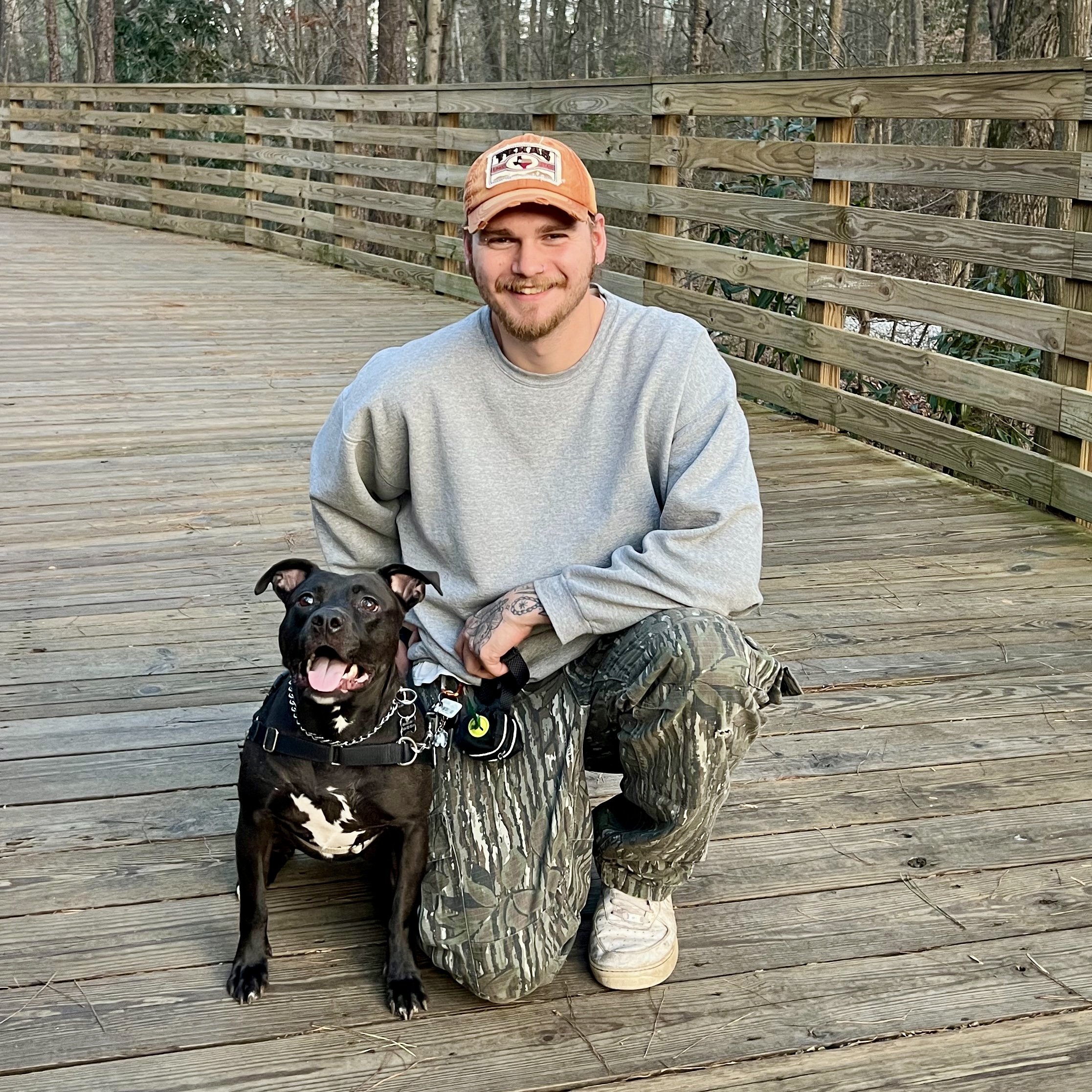 A young man kneeling on a wooden bridge outdoors with a smiling black dog that has white markings, wearing a harness and a chain collar, in a natural wooded setting.